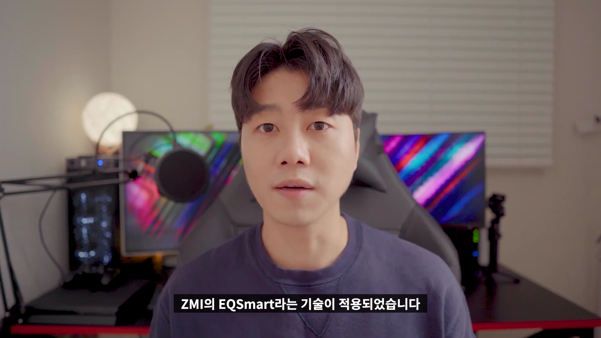 실내에서 카메라를 응시하며 말하는 남성 뒤로 모니터와 조명이 배치된 책상이 보인다.