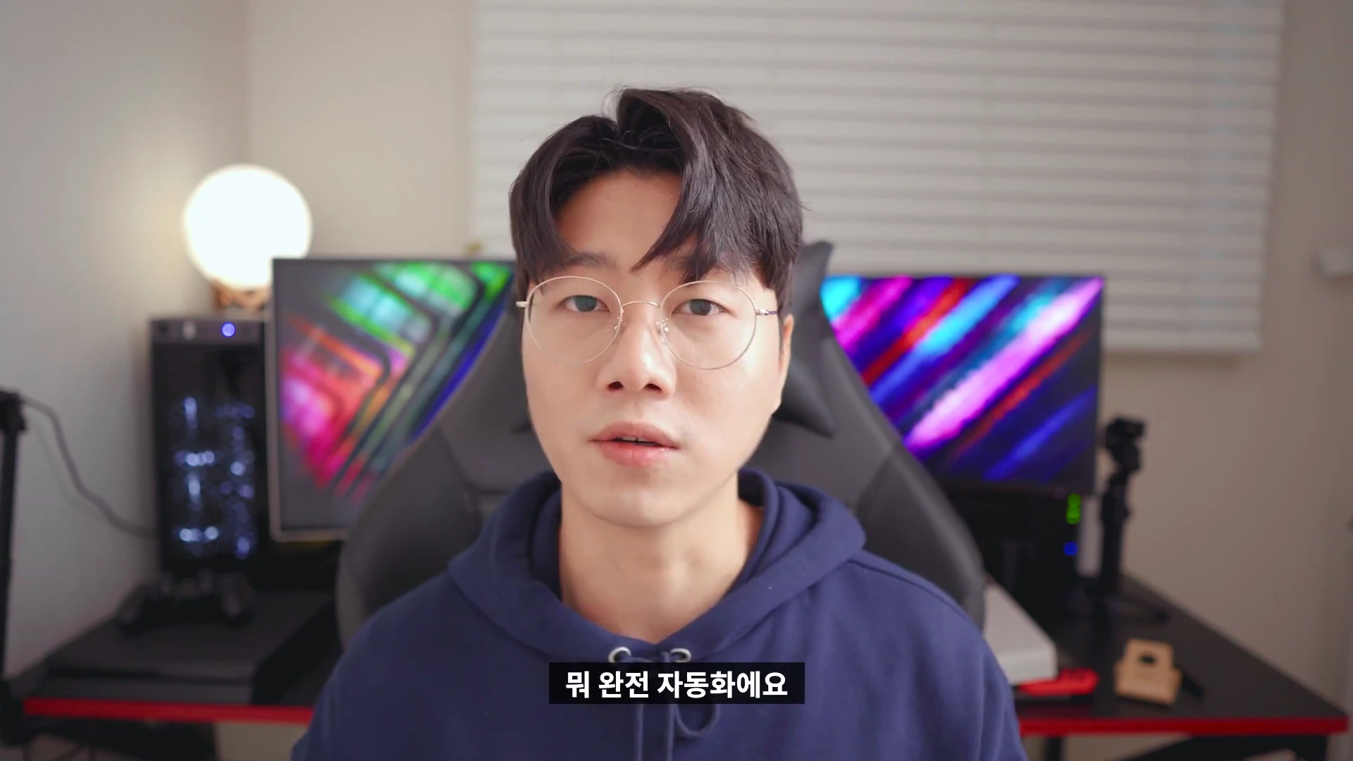 안경을 쓴 남성이 책상 앞에 앉아 카메라를 정면으로 응시하고 있는 모습
