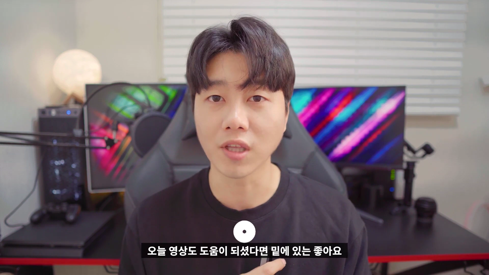 검은색 상의를 입은 남성이 책상 앞에 앉아 카메라를 정면으로 응시하며 이야기하고 있다.