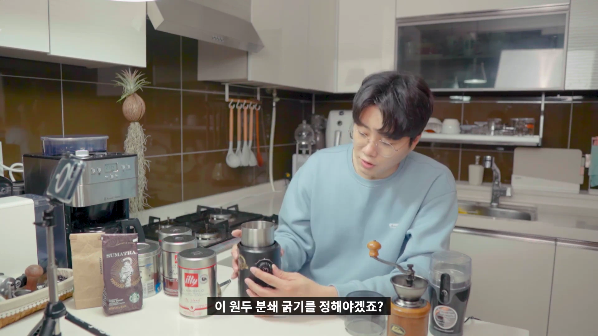 주방에서 안경을 쓴 남성이 전동 그라인더의 다이얼을 조절하며 원두 분쇄 굵기를 설정하고 있다.