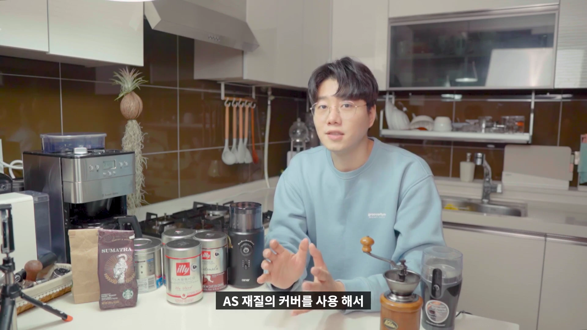 주방에서 안경을 쓴 남성이 원두 그라인더 제품을 앞에 두고 설명하고 있다.