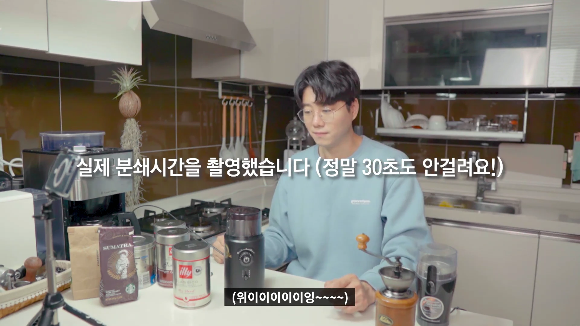 주방 테이블에 앉아 전동 그라인더를 사용하는 남성과 주변에 놓인 원두 패키지들