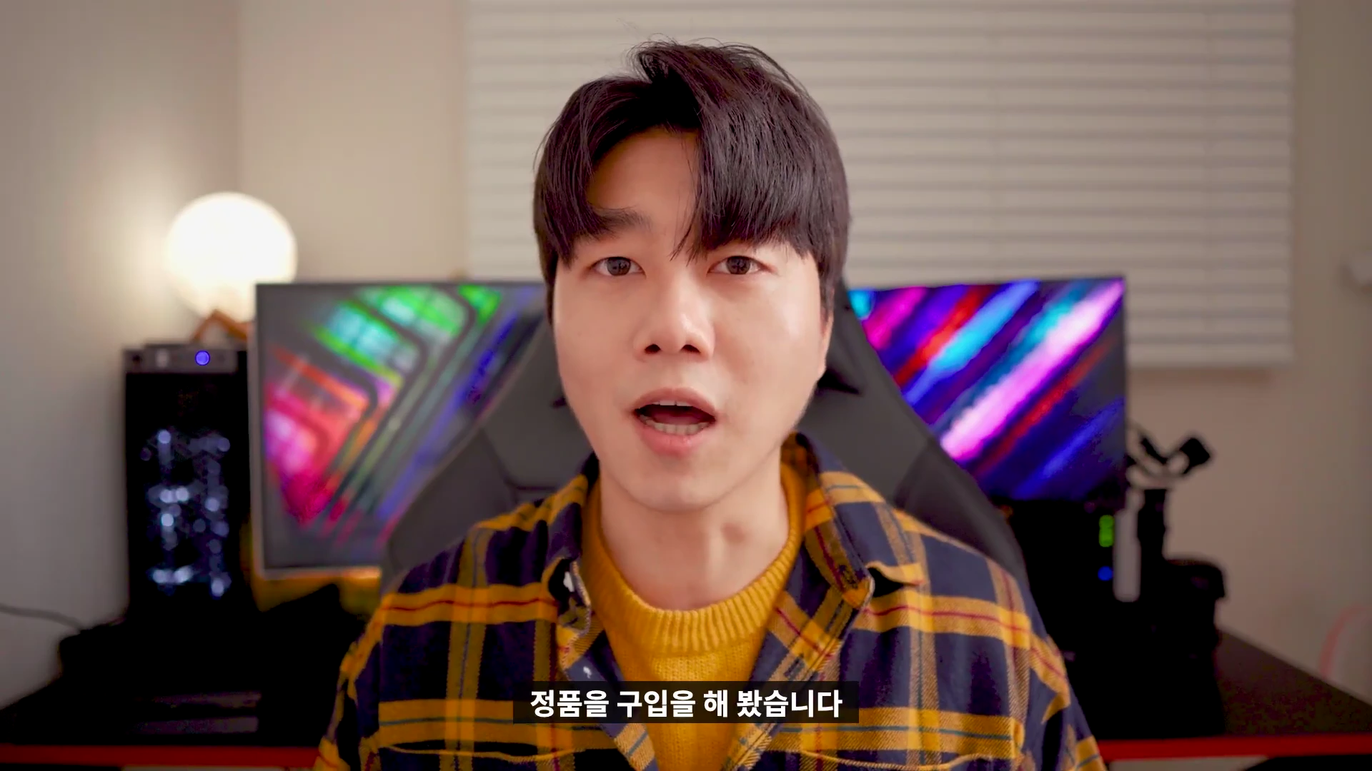 체크무늬 셔츠를 입은 남성이 실내에서 카메라를 정면으로 응시하며 이야기하고 있다.