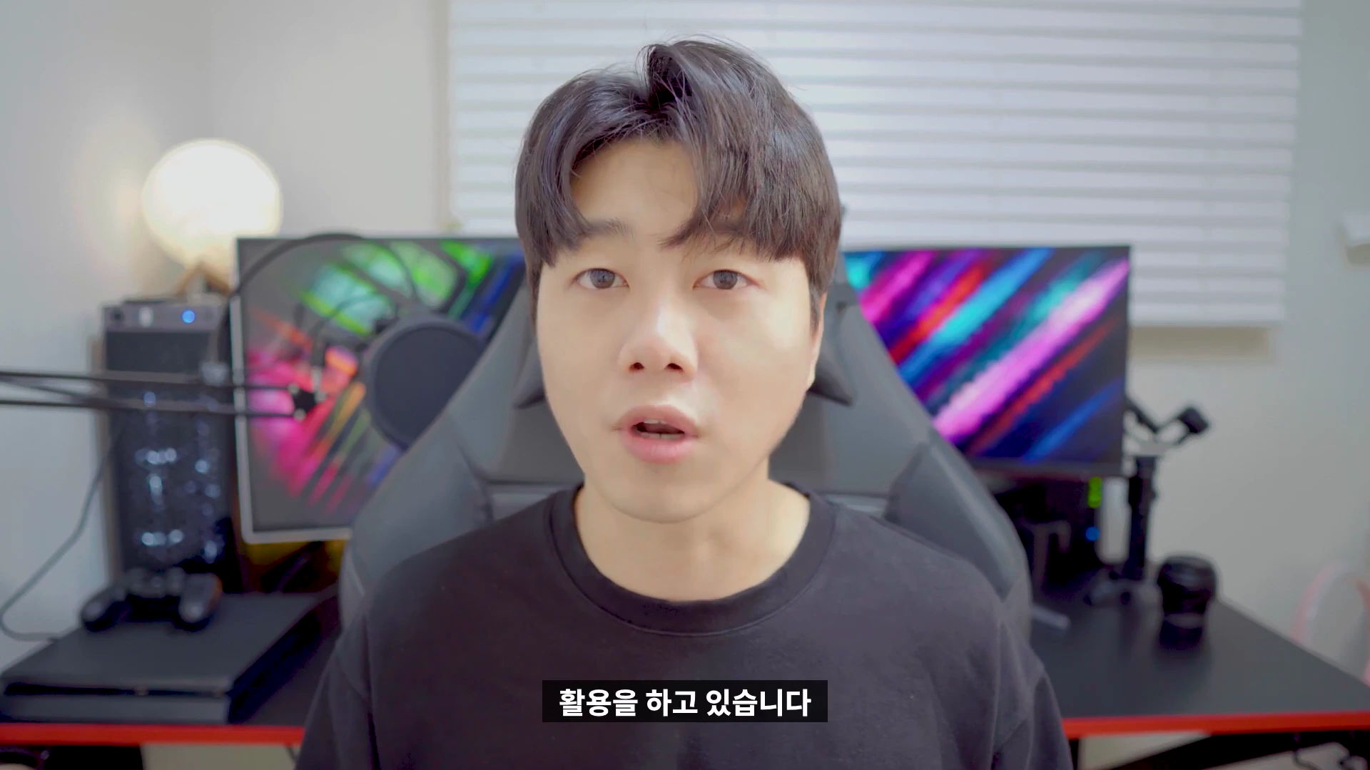검은색 티셔츠를 입은 남성이 실내 책상 앞에 앉아 정면을 바라보며 이야기하고 있다.