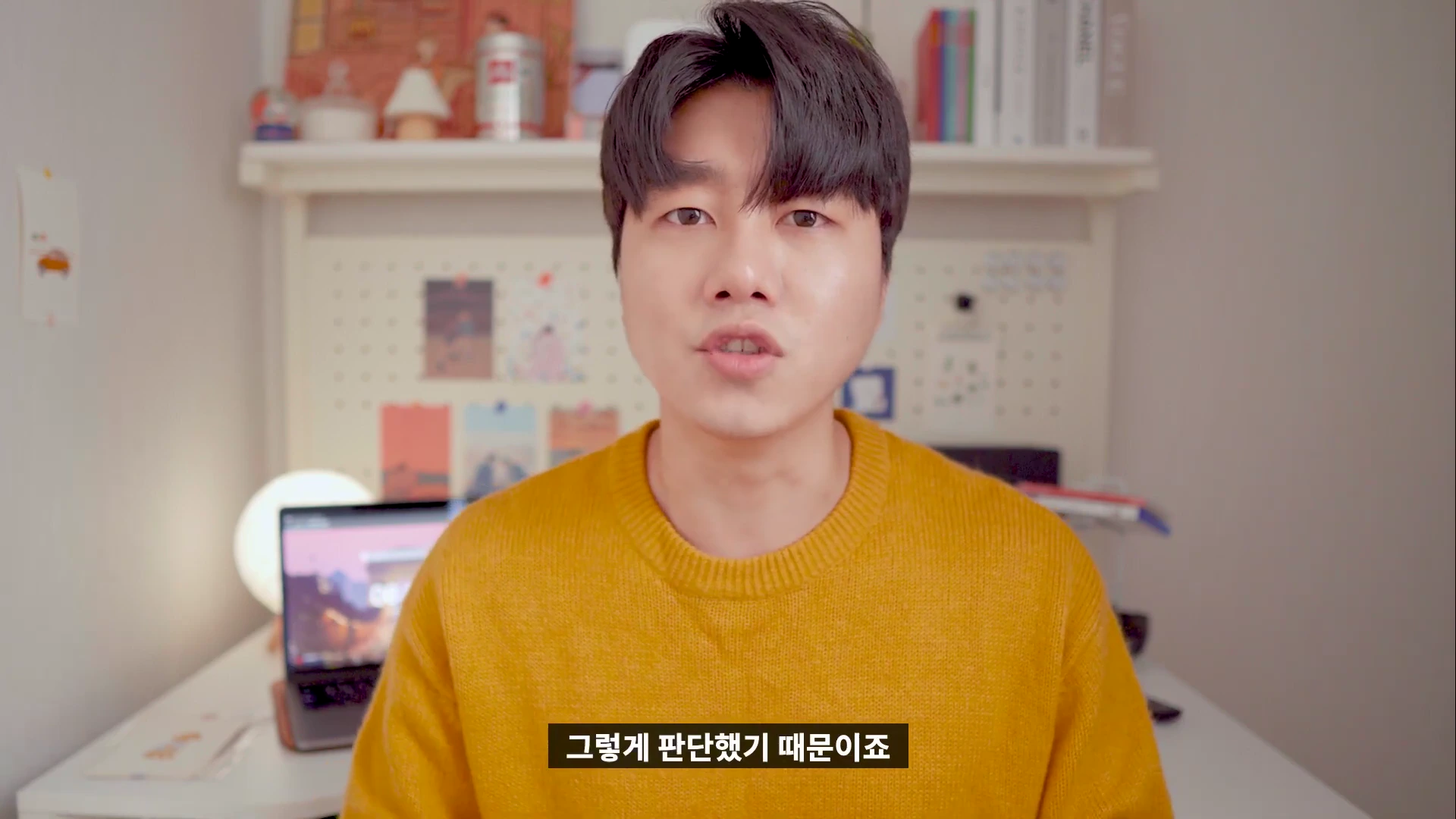 노란색 니트를 입은 남성이 책상 앞에 앉아 정면을 바라보며 이야기하는 모습