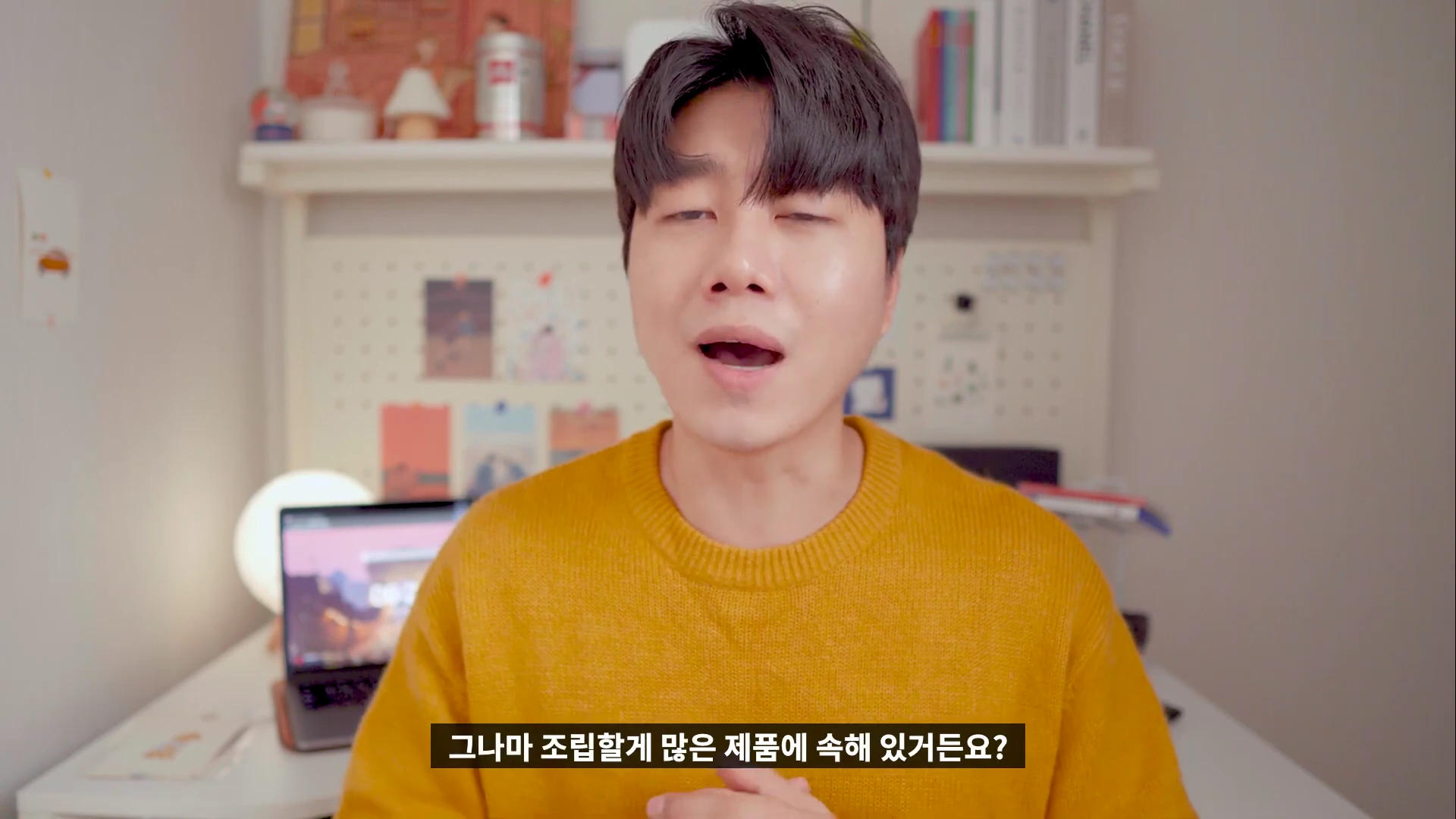 노란색 니트를 입은 남성이 실내에서 카메라를 보며 이야기하고 있다.