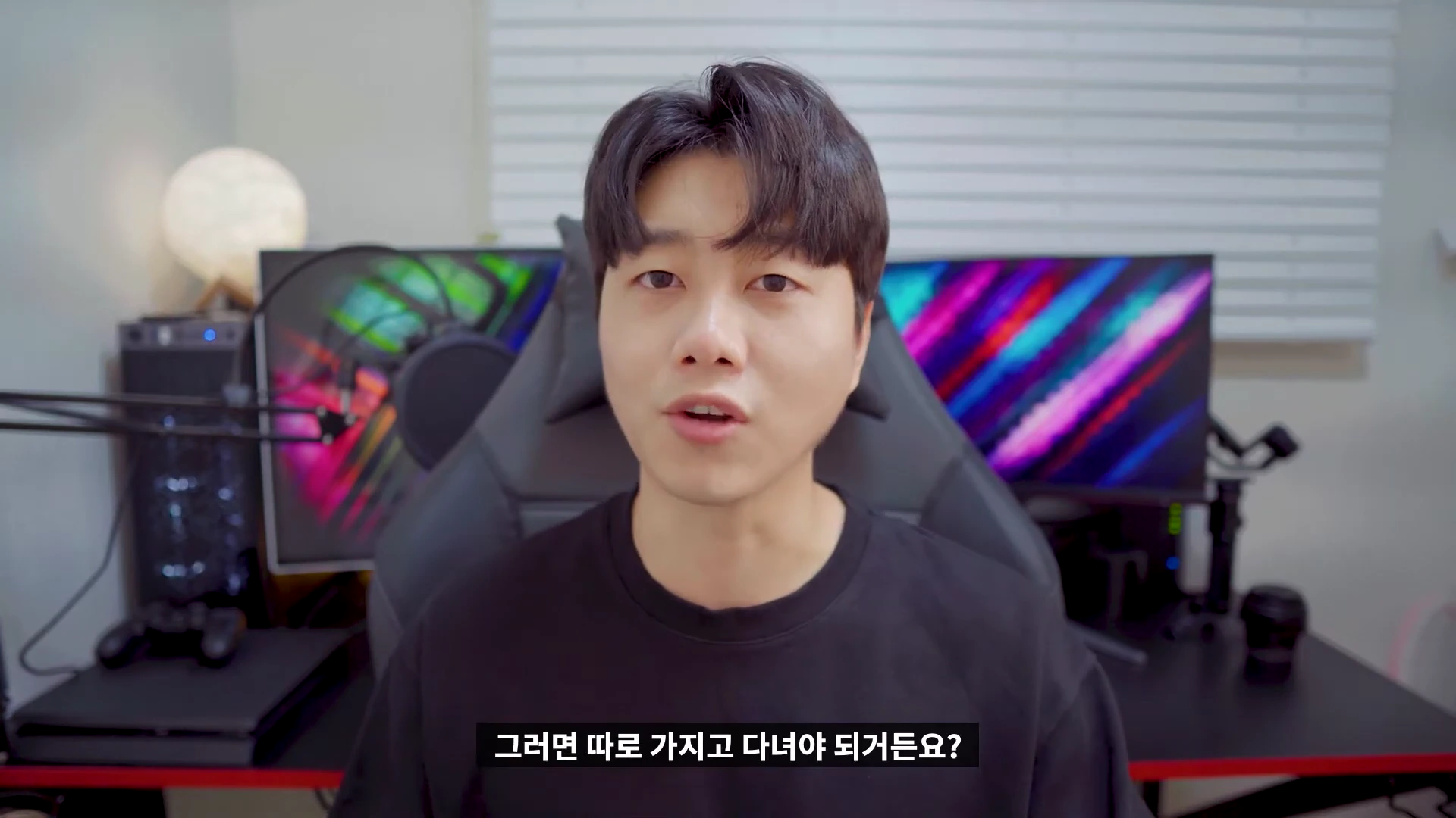 검은색 티셔츠를 입은 남성이 책상 앞에 앉아 카메라를 보며 이야기하는 모습