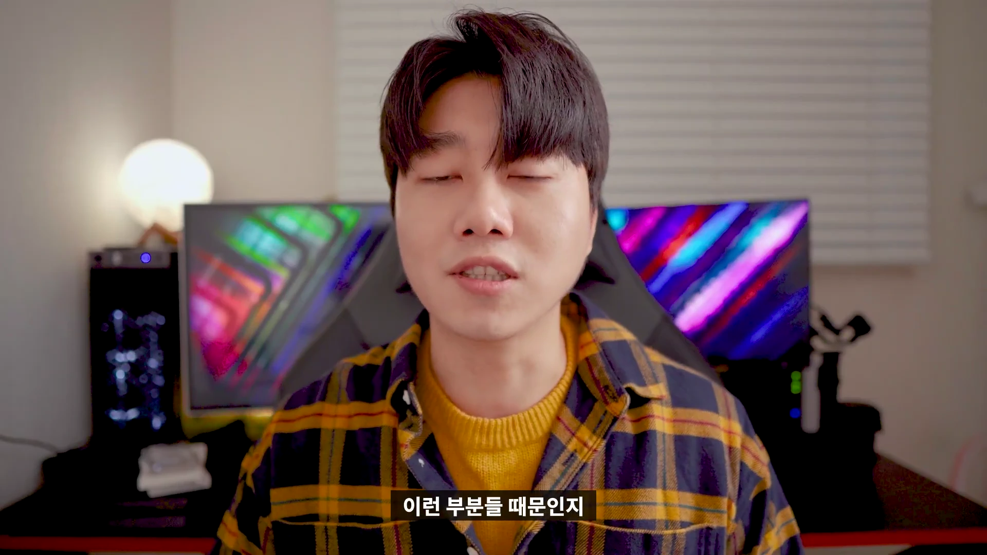 체크무늬 셔츠를 입은 남성이 실내에서 카메라를 보며 이야기하고 있는 모습