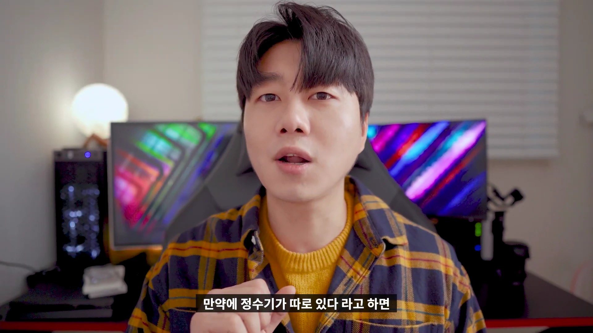 체크무늬 셔츠를 입은 남성이 실내에서 카메라를 응시하며 이야기를 나누고 있다.