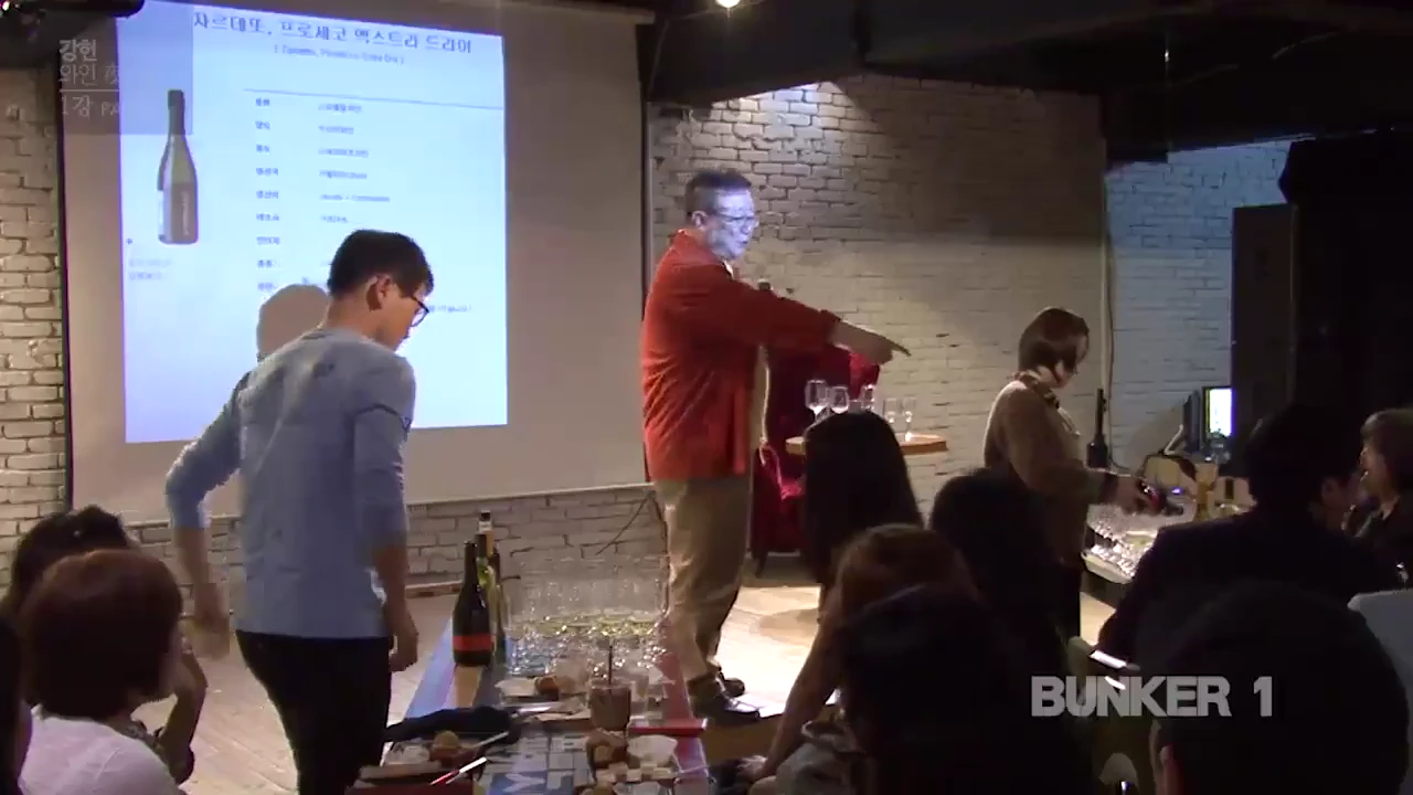 강연장에서 빨간 셔츠를 입은 강사가 청중에게 와인 테이스팅 방법을 설명하고 있으며, 뒤쪽 스크린에는 와인 정보가 담긴 슬라이드가 띄워져 있다.