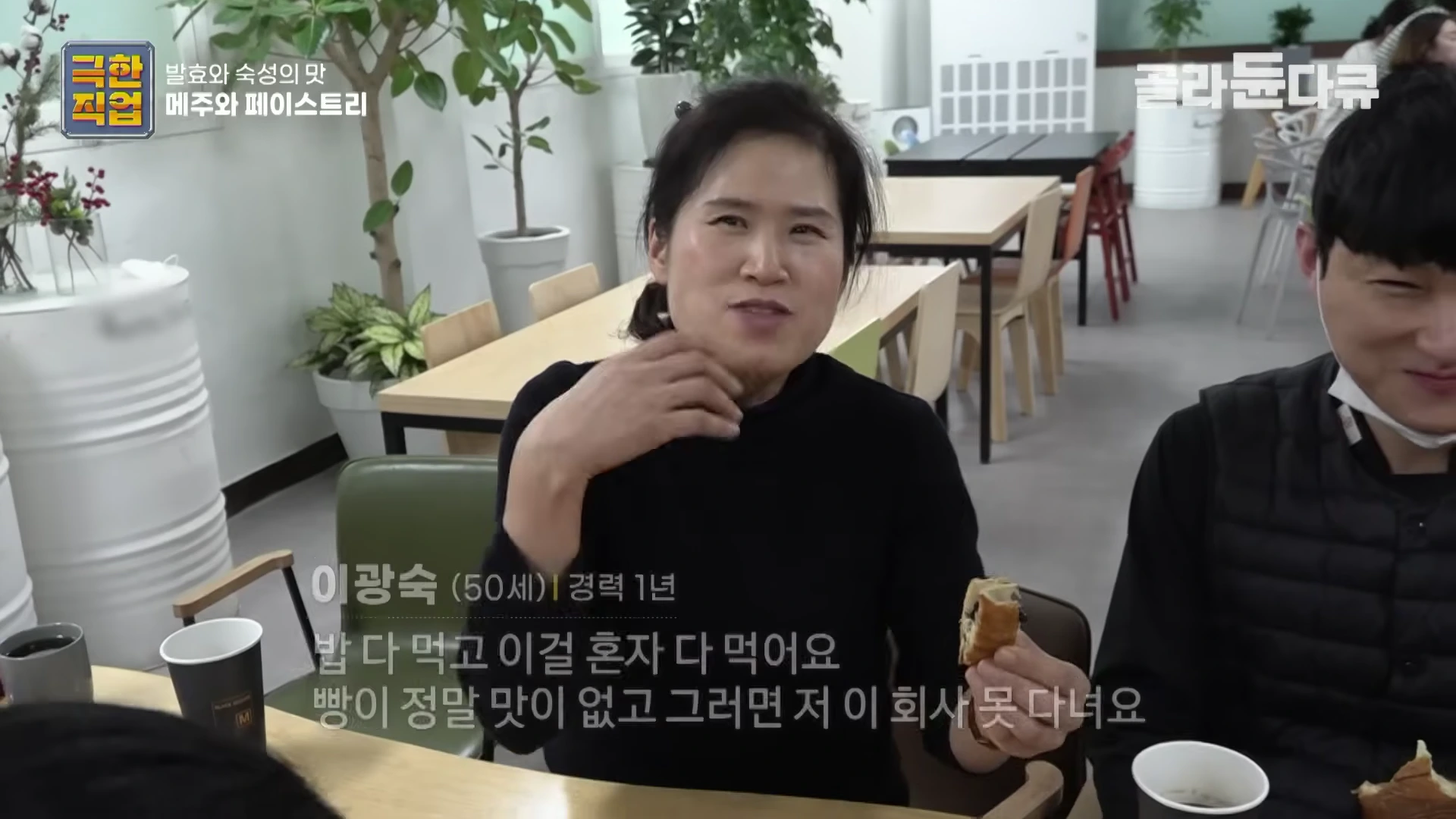 실내에서 검은 옷을 입은 중년 여성이 크루아상을 손에 들고 동료들과 함께 간식을 먹으며 인터뷰를 하고 있다.