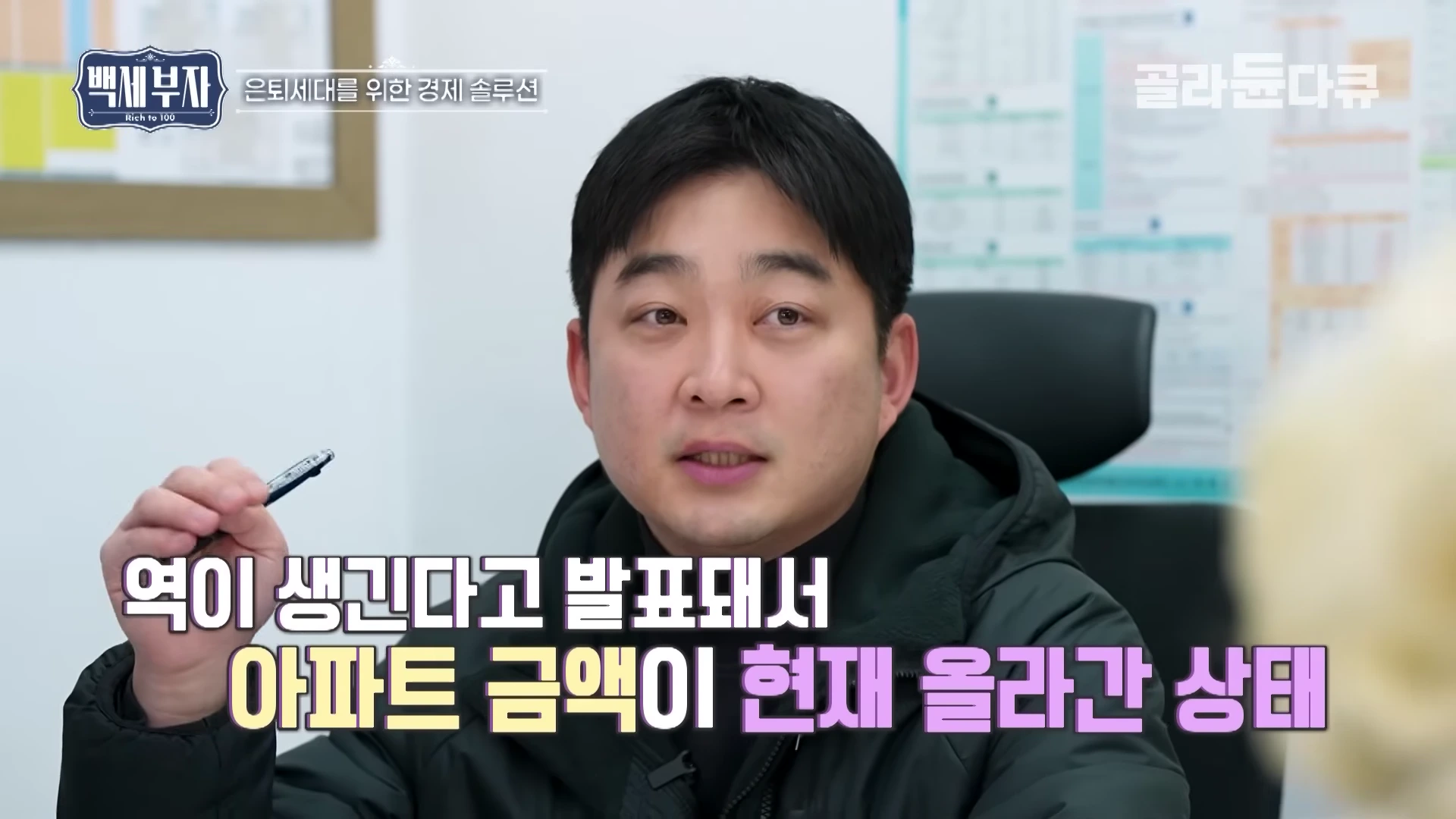 실내에서 펜을 든 남성이 대화하며 설명하는 모습과 '역이 생긴다고 발표돼서 아파트 금액이 현재 올라간 상태'라는 자막이 하단에 표시된 영상 화면