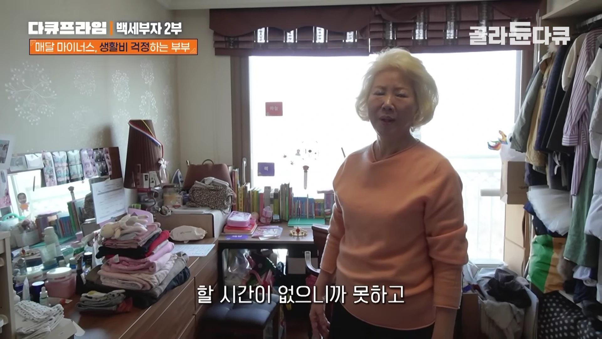 밝은색 티셔츠를 입은 중년 여성이 거실 가득 짐과 옷이 가득한 집 안에서 서 있는 모습