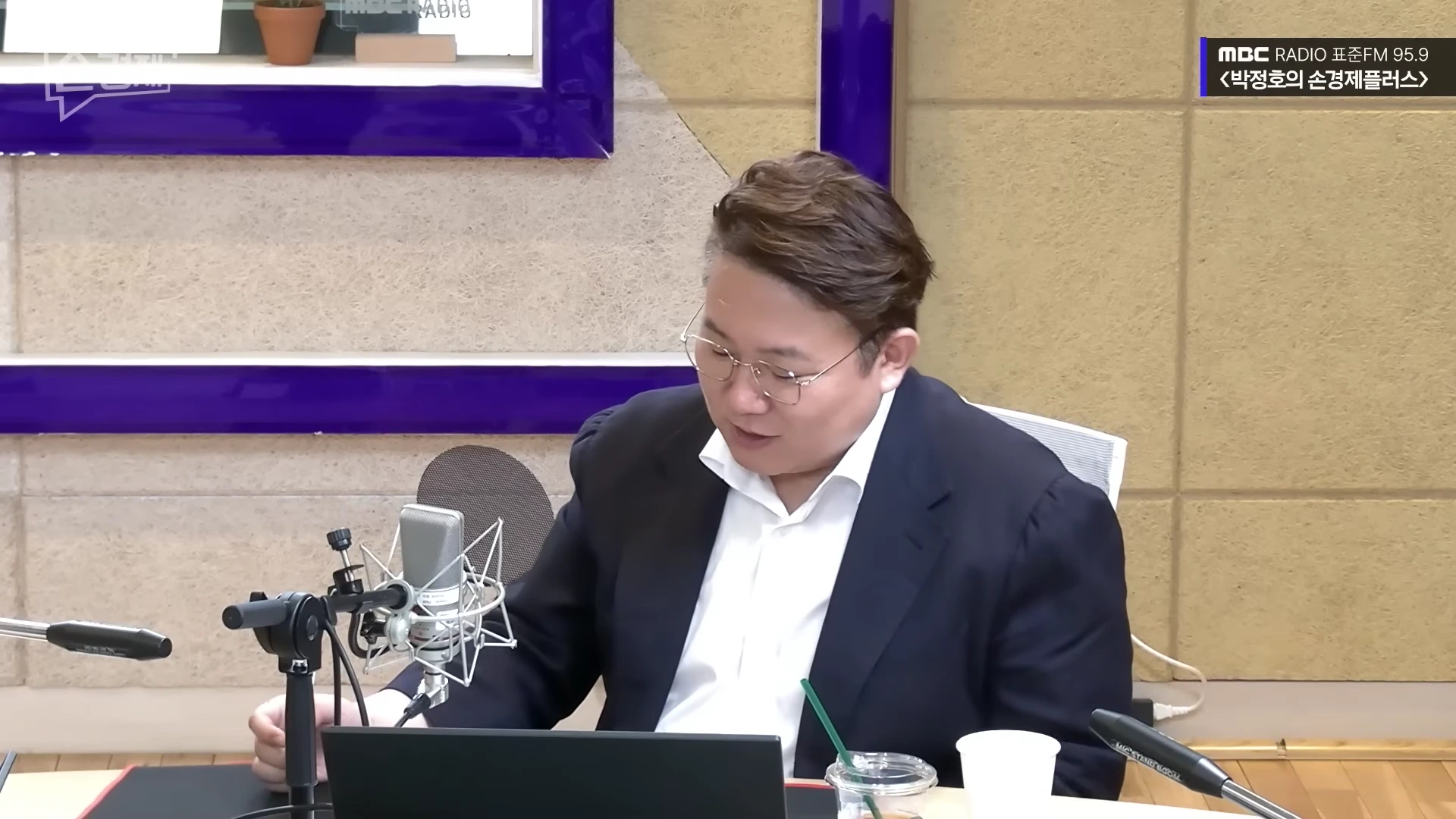 안경을 쓴 남성이 라디오 스튜디오 마이크 앞에 앉아 대화를 나누는 모습