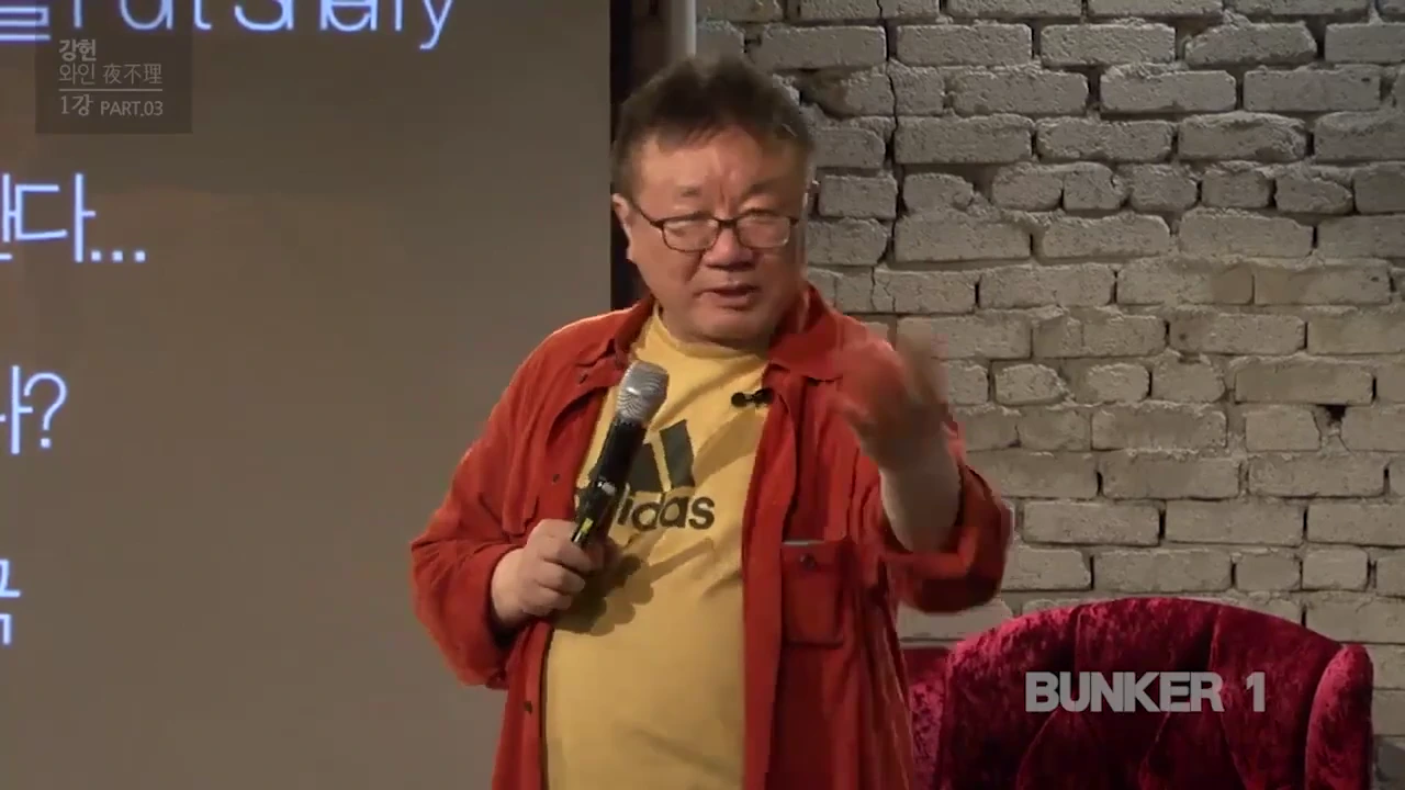 안경을 쓴 중년 남성이 노란색 티셔츠와 붉은 셔츠를 입고 마이크를 든 채 이야기를 나누는 모습