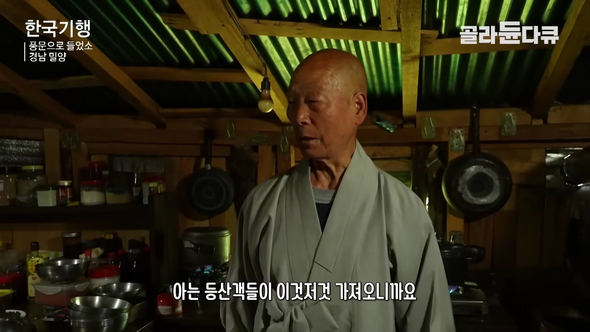 회색 승복을 입은 스님이 나무 벽으로 둘러싸인 주방에서 등산객들이 가져온 물건들에 대해 이야기하고 있다.