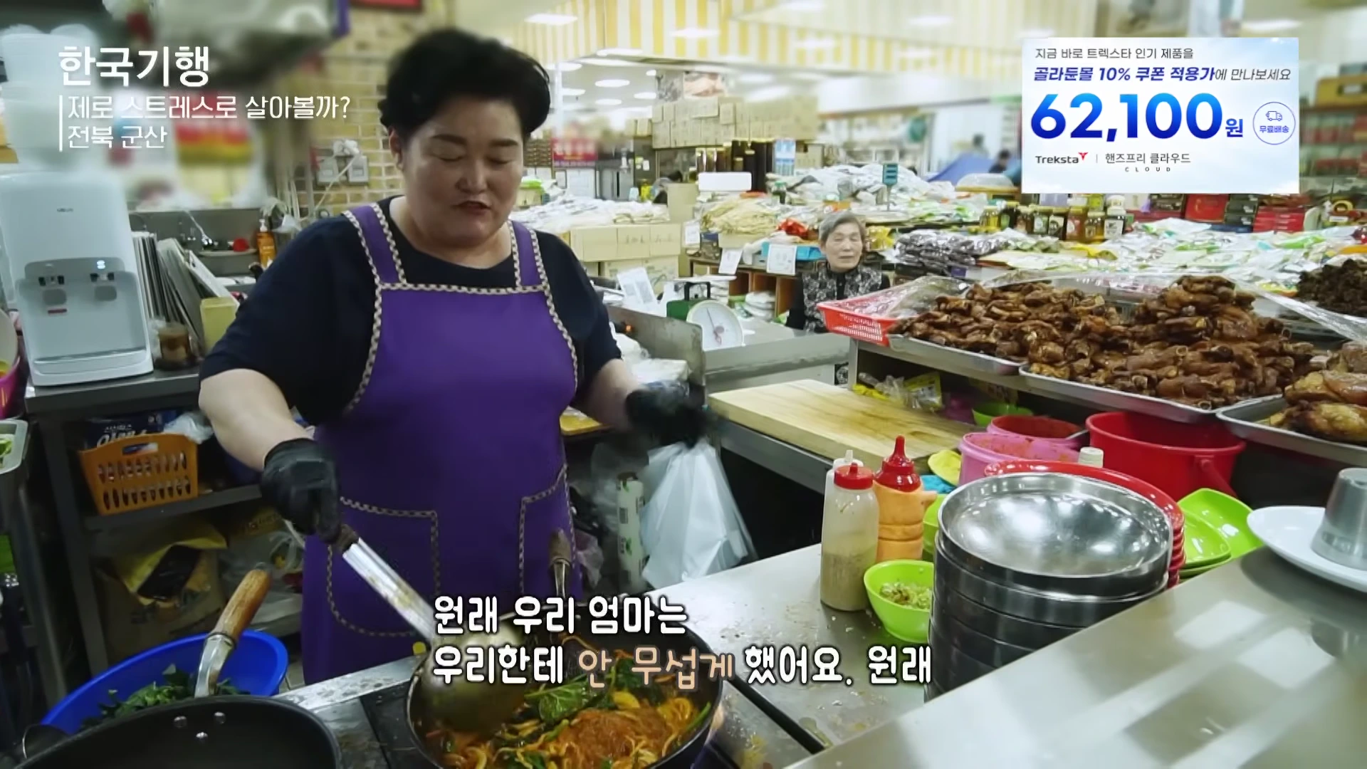 보라색 앞치마를 두른 여성이 시장의 좁은 조리 공간에서 프라이팬에 잡채를 볶고 있는 모습