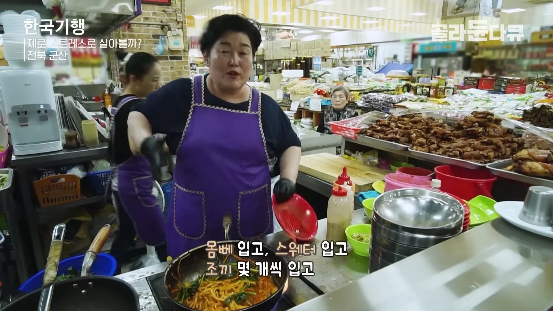 보라색 앞치마를 두른 여성이 시장 상가 주방에서 프라이팬으로 조리하며 앞에 서 있는 모습