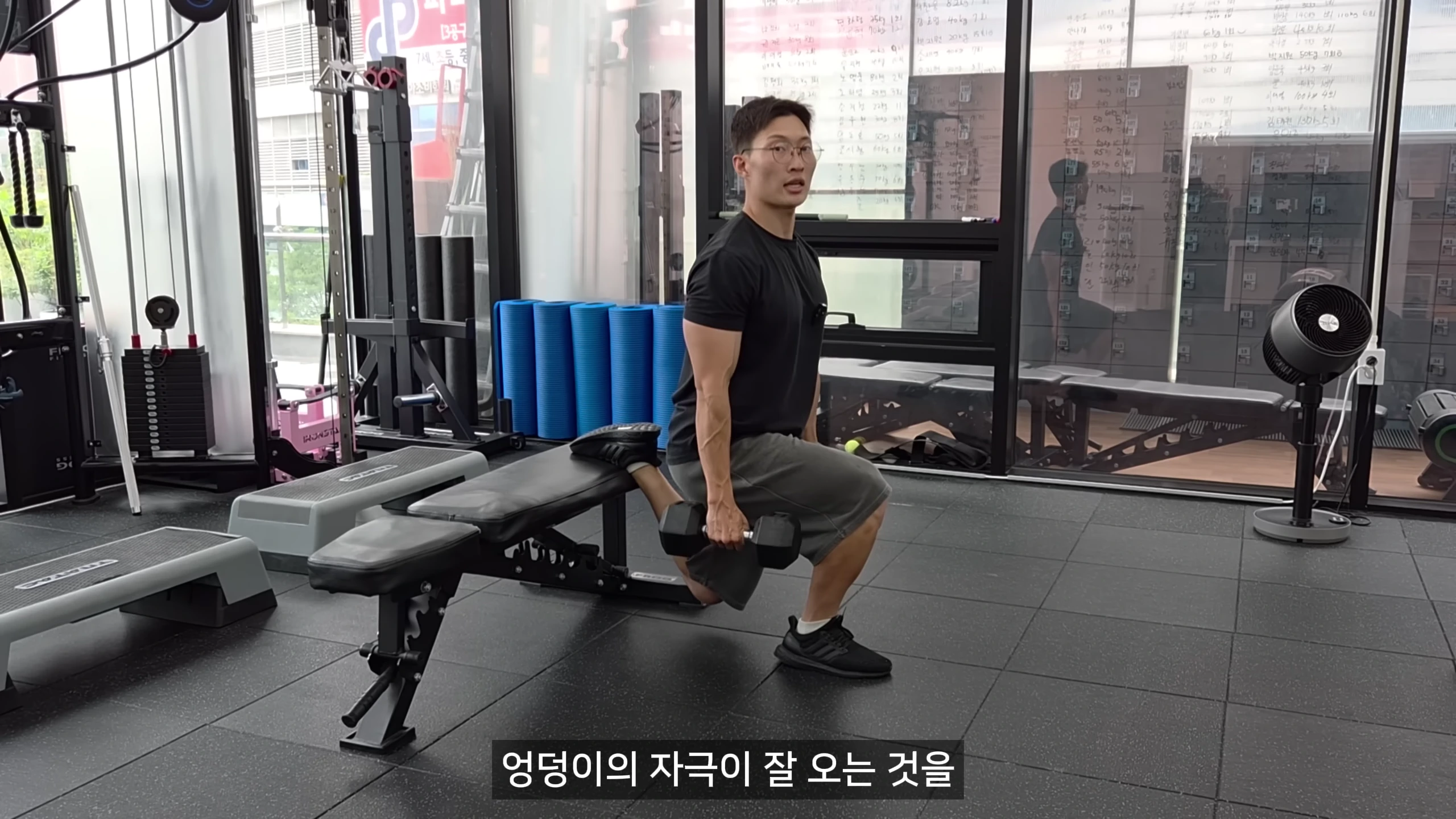 체육관에서 덤벨을 들고 한쪽 발을 벤치에 올린 채 불가리안 스플릿 스쿼트 자세를 취하고 있는 남성