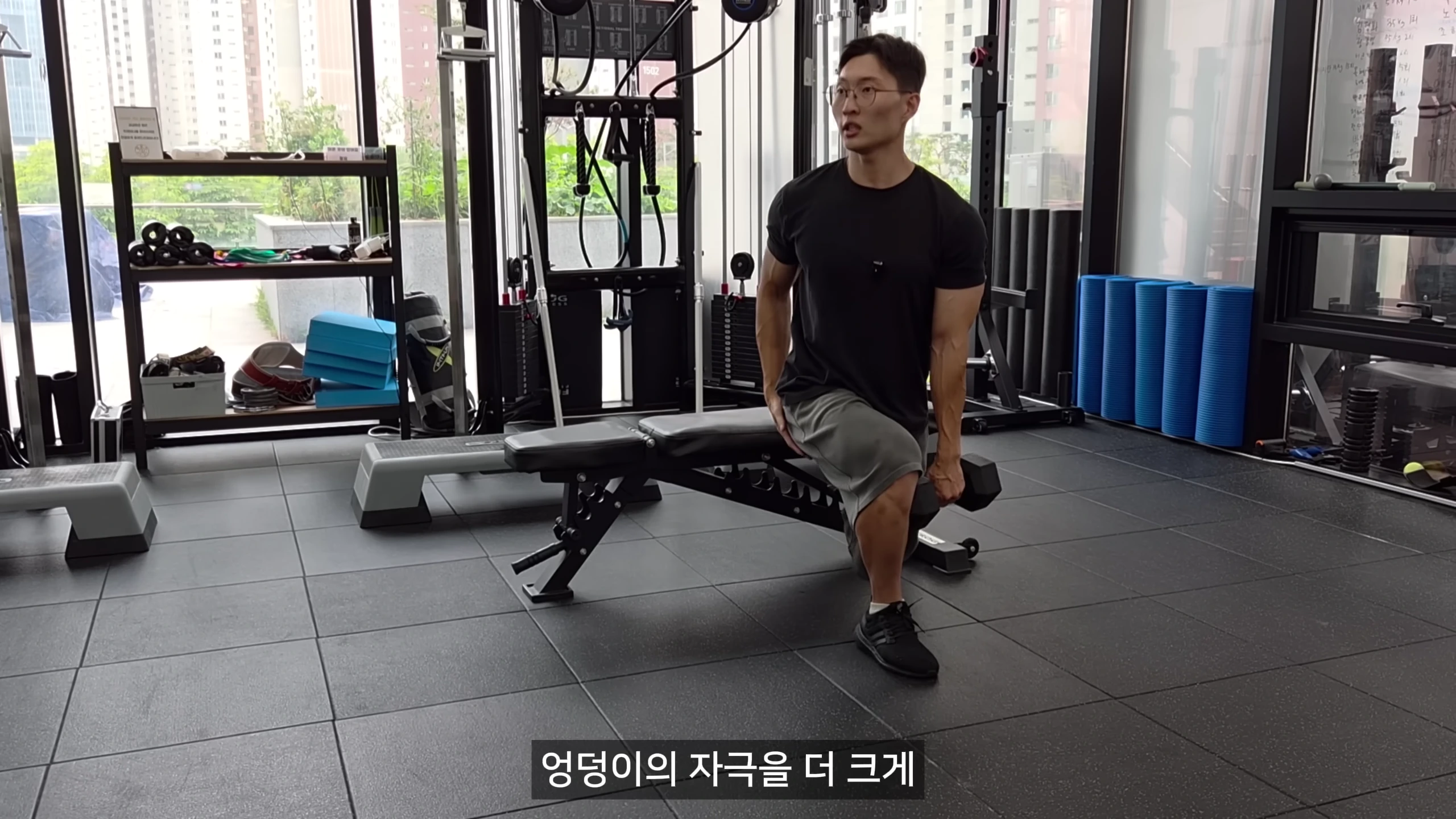헬스장에서 검은색 티셔츠를 입은 남성이 벤치에 한쪽 다리를 올리고 덤벨을 든 채 스쿼트 자세를 연습하고 있다.