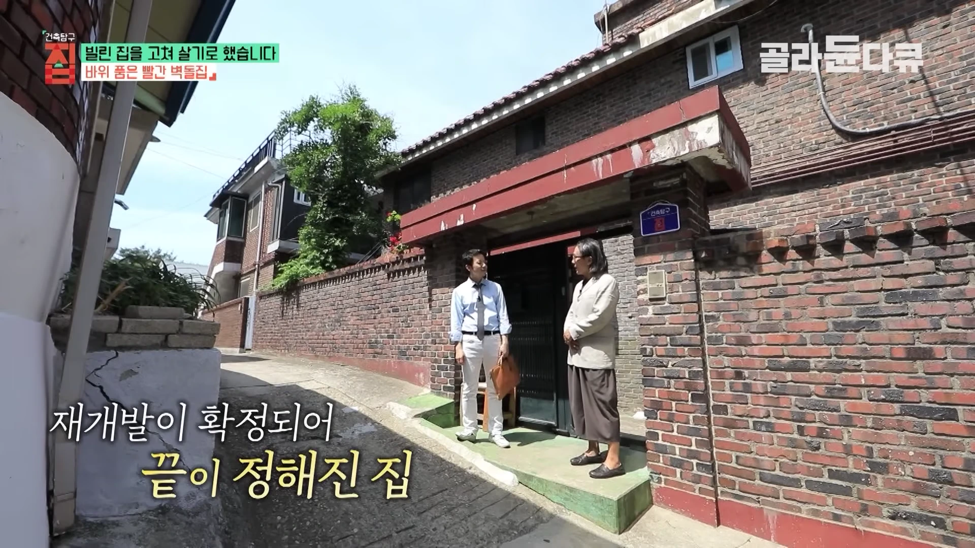 두 남자가 신당동 언덕길에 있는 낡은 빨간 벽돌집 앞에서 대화를 나누고 있다.