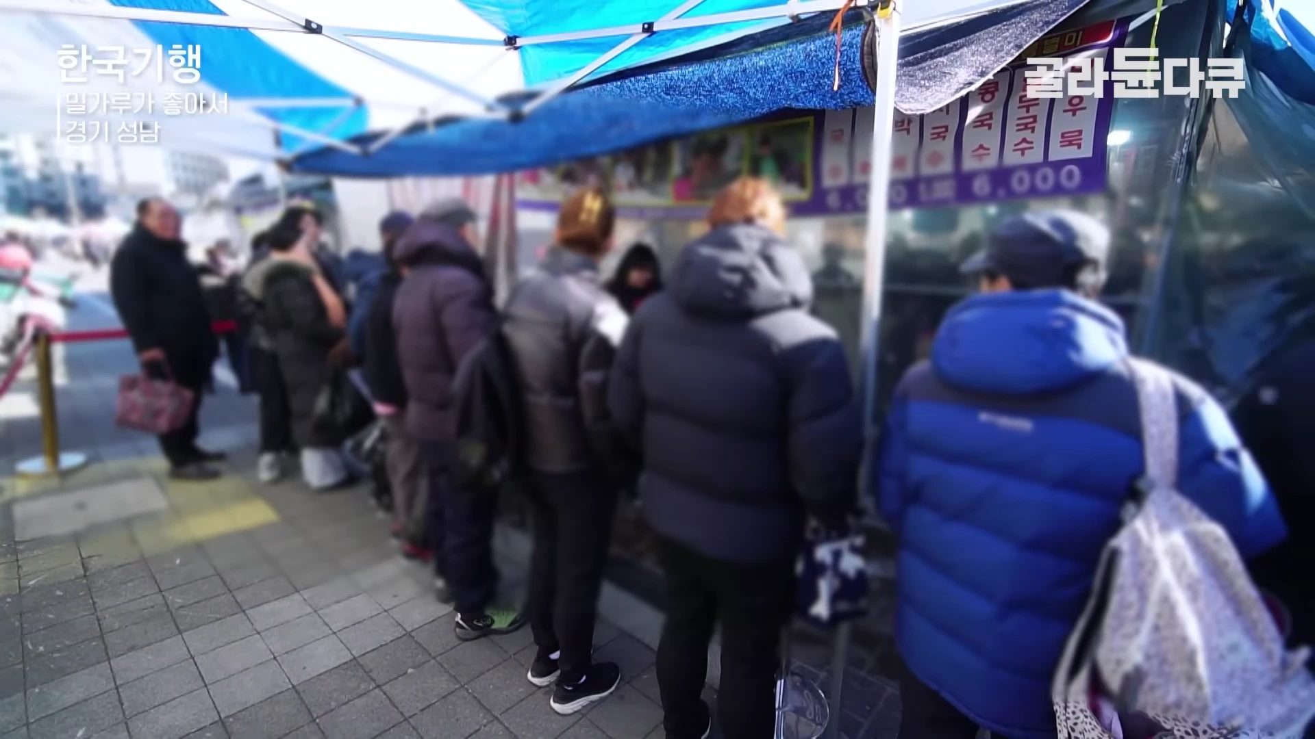 겨울 외투를 입은 사람들이 야외 시장 가판대 앞에서 음식을 사기 위해 길게 줄을 서 있다.