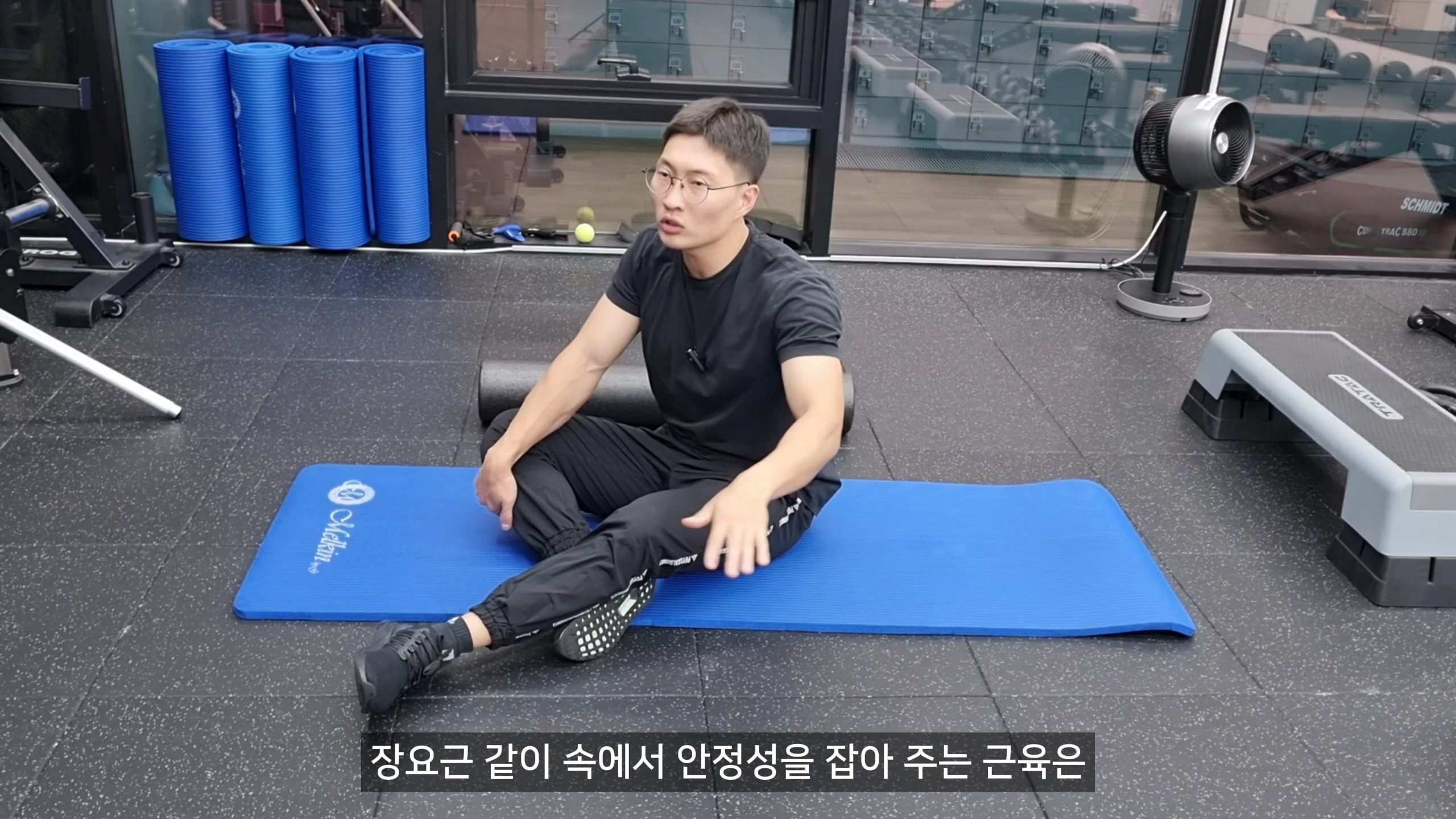 운동복 차림의 남성이 파란색 매트 위에서 앉아 손동작을 섞어 운동 원리를 설명하고 있다.