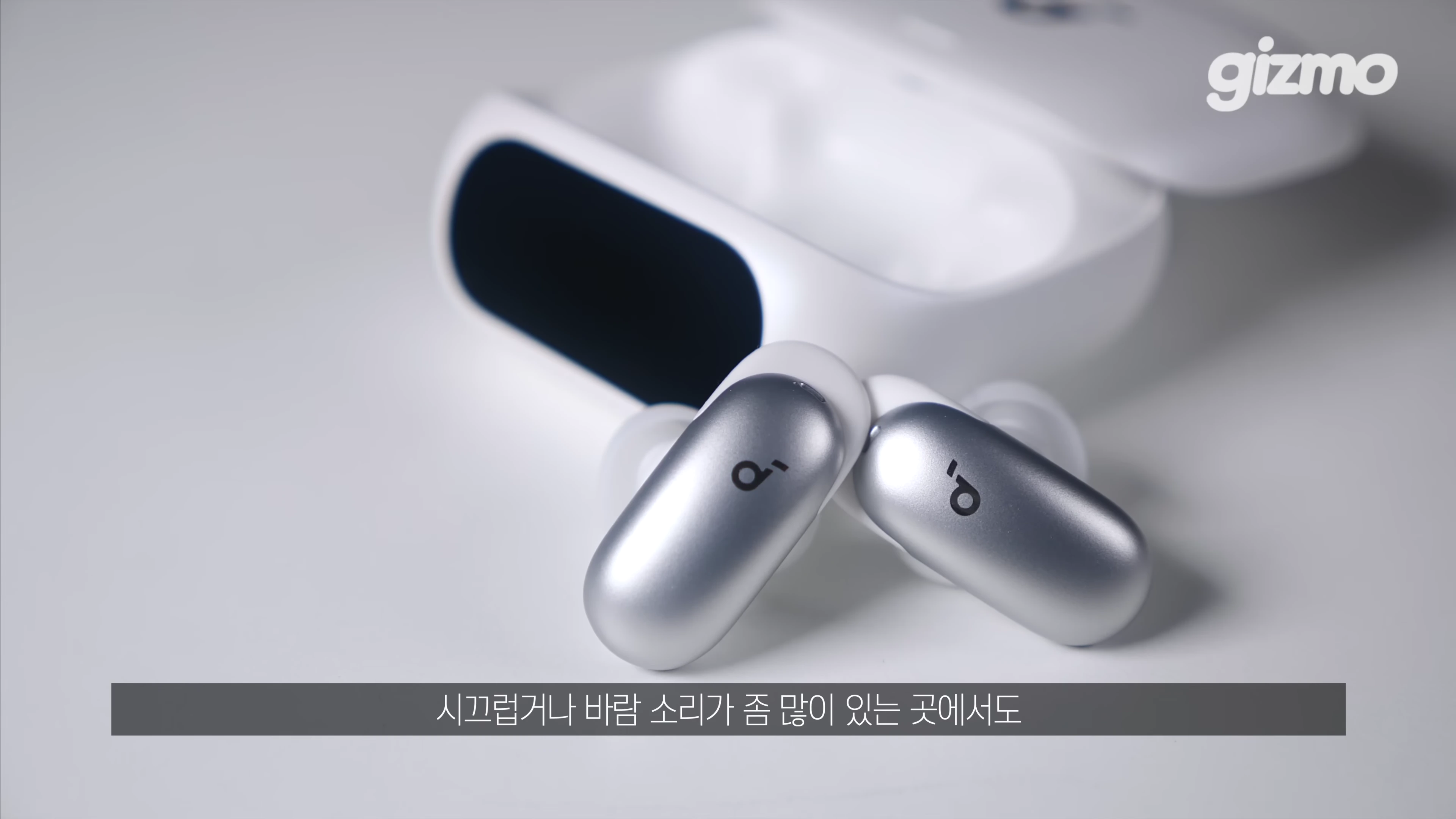 흰색 무선 이어폰 충전 케이스와 그 앞에 놓인 메탈릭 실버 색상의 이어폰 유닛 한 쌍이 밝은 배경 위에 놓여 있다.