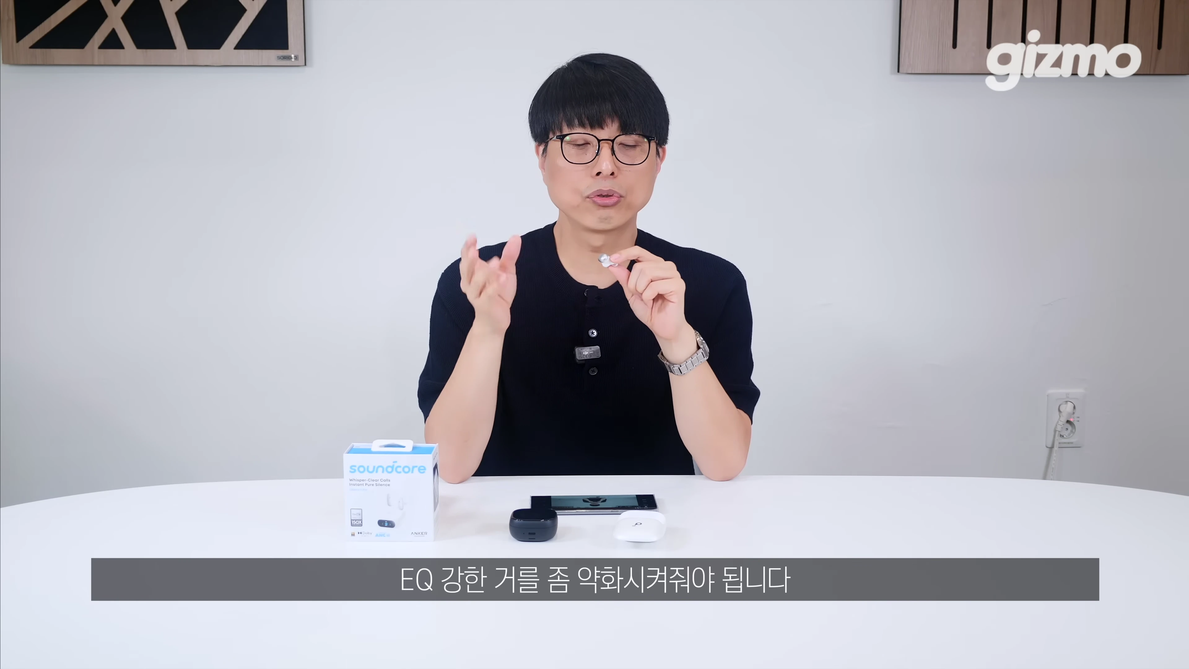안경을 쓴 남성이 흰색 테이블에 무선 이어폰 케이스와 패키지 박스를 두고 제품을 직접 들어 보이며 설명하고 있다.