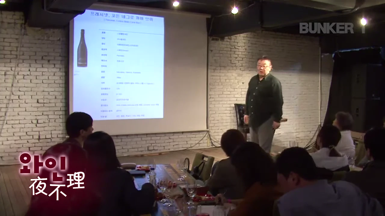 강연자가 스크린에 띄워진 특정 와인 정보를 가리키며 청중 앞에서 와인 강의를 하고 있다.