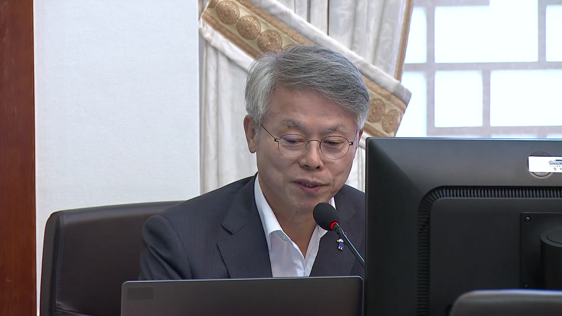 정장을 입고 안경을 쓴 중년 남성이 회의실 책상에 앉아 마이크 앞에 서서 이야기를 나누고 있다.