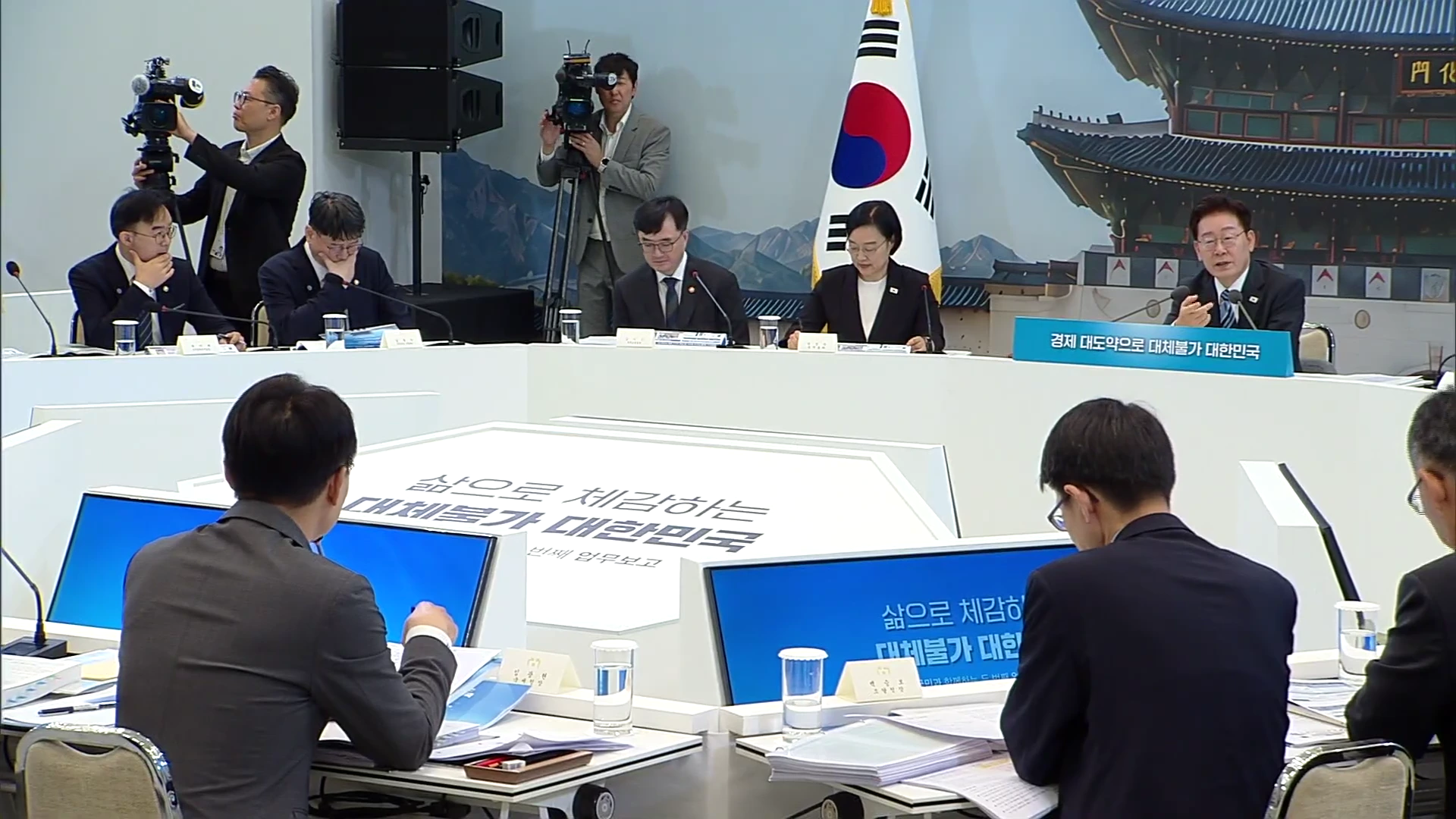 회의장에 여러 명의 관계자가 앉아 있고 한 사람이 마이크를 잡고 발표를 하는 가운데 카메라 기자가 촬영 중인 모습