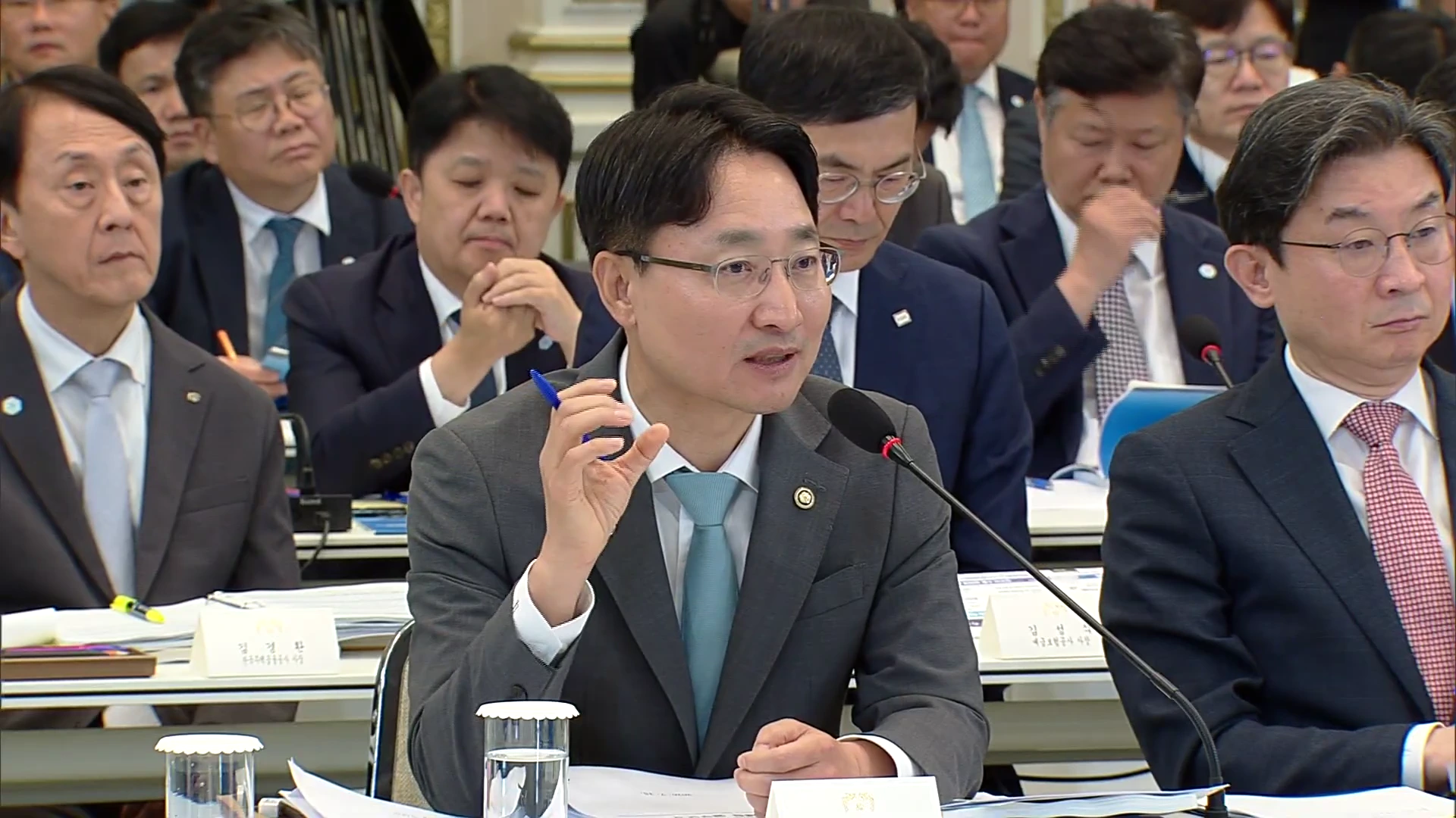 정장을 차려입은 국세청장이 회의장에서 마이크를 잡고 손동작을 하며 발언하고 있습니다. 주변에는 정장을 입은 관계자들이 진지한 표정으로 회의에 집중하고 있습니다.