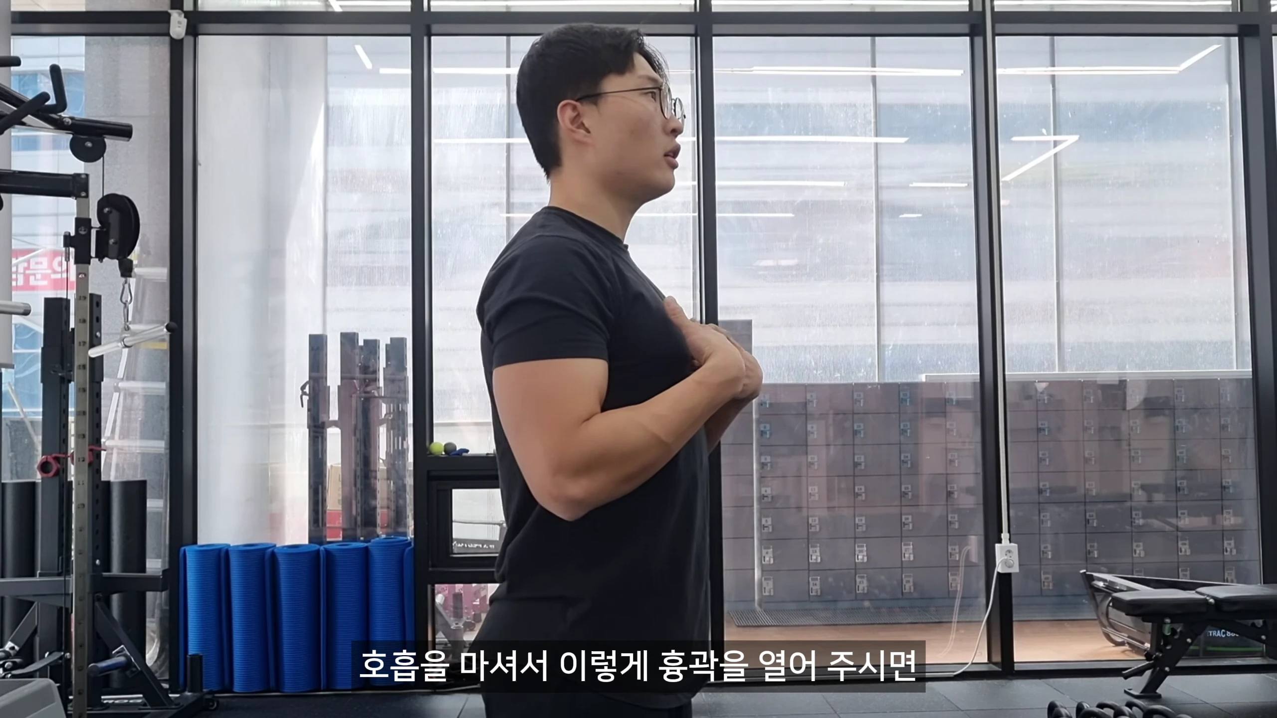 체육관에서 검은색 티셔츠를 입은 남성이 서서 호흡으로 흉곽을 확장하는 동작을 보여주고 있는 모습.