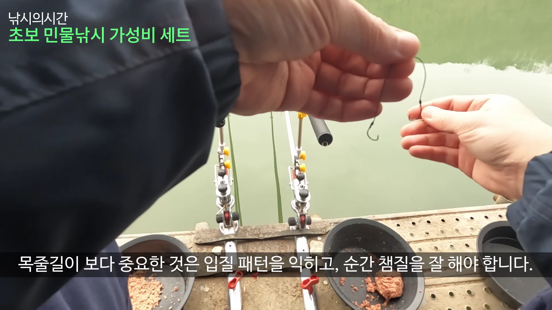낚시인이 강가에서 손으로 낚싯바늘과 줄을 잡고 있고, 앞에는 낚싯대 두 대가 받침대에 놓여 있으며 떡밥 그릇이 보입니다.
