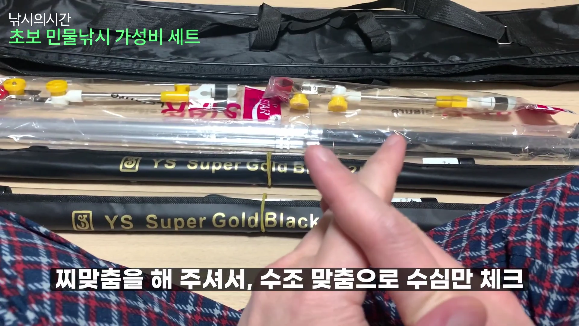 바닥에 놓인 민물낚시 세트 구성품과 검은색 낚시 가방, 그리고 그 앞에서 손가락 하트를 만들고 있는 사람의 손.