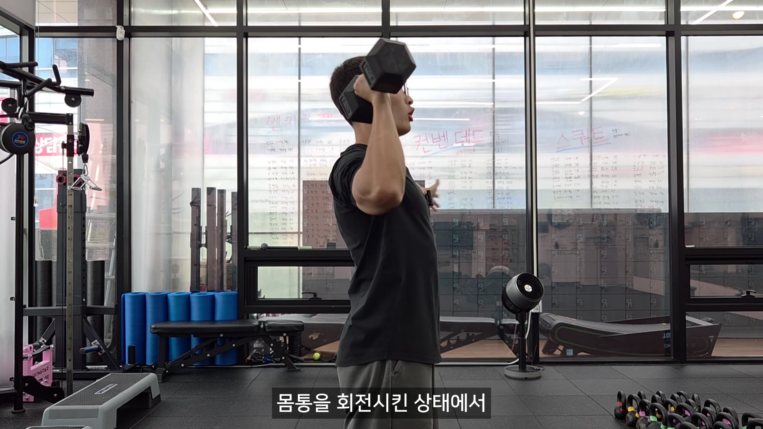 체육관에서 검정색 티셔츠를 입은 남성이 덤벨을 들고 숄더 프레스 동작을 하고 있는 옆모습