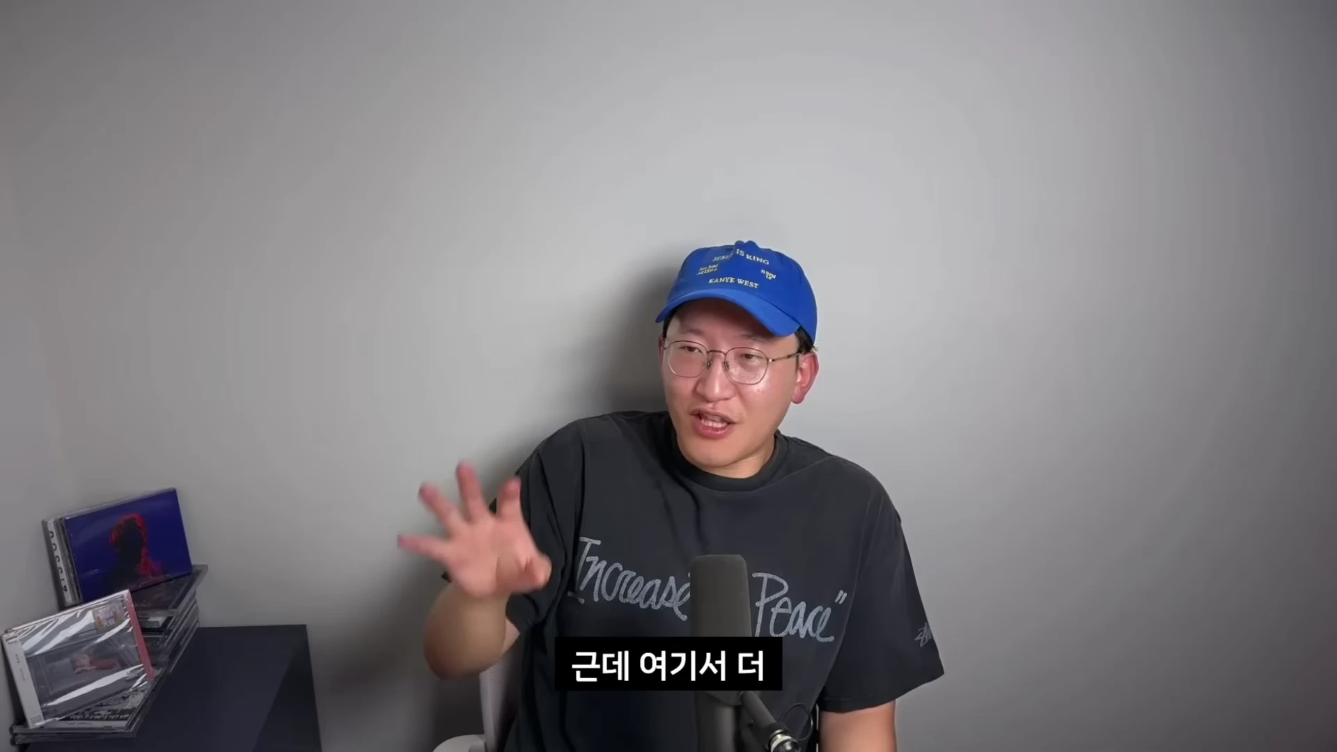 안경을 쓰고 파란색 야구 모자를 쓴 남성이 마이크 앞에 앉아 손을 들어 올리며 이야기를 나누고 있다.