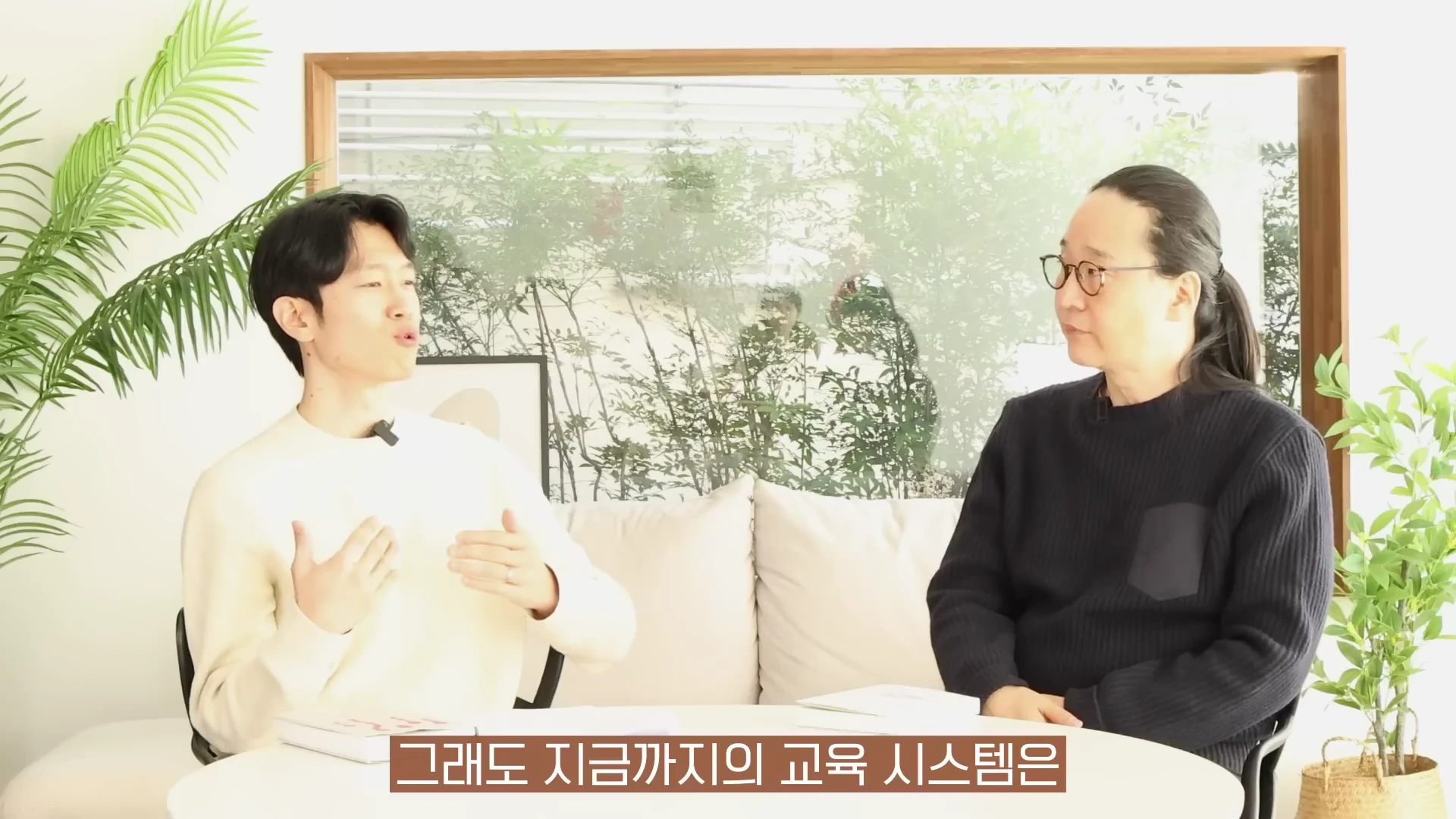 밝은 햇살이 들어오는 창가 테이블에 마주 앉아 대화를 나누고 있는 두 사람의 모습이며, 아래쪽에는 한국어 자막이 노란색 박스 안에 표시되어 있습니다.