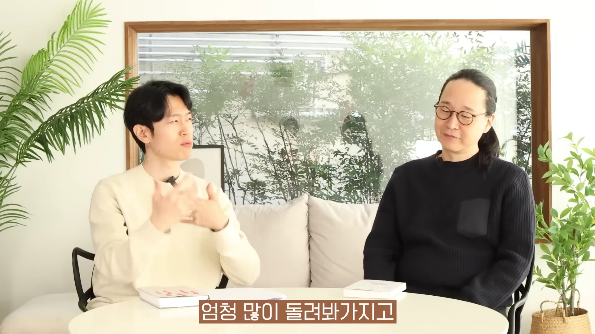 밝은 실내에서 두 남성이 흰색 원형 테이블을 사이에 두고 마주 앉아 대화하는 모습이며 테이블 위에는 책 몇 권이 놓여 있습니다.