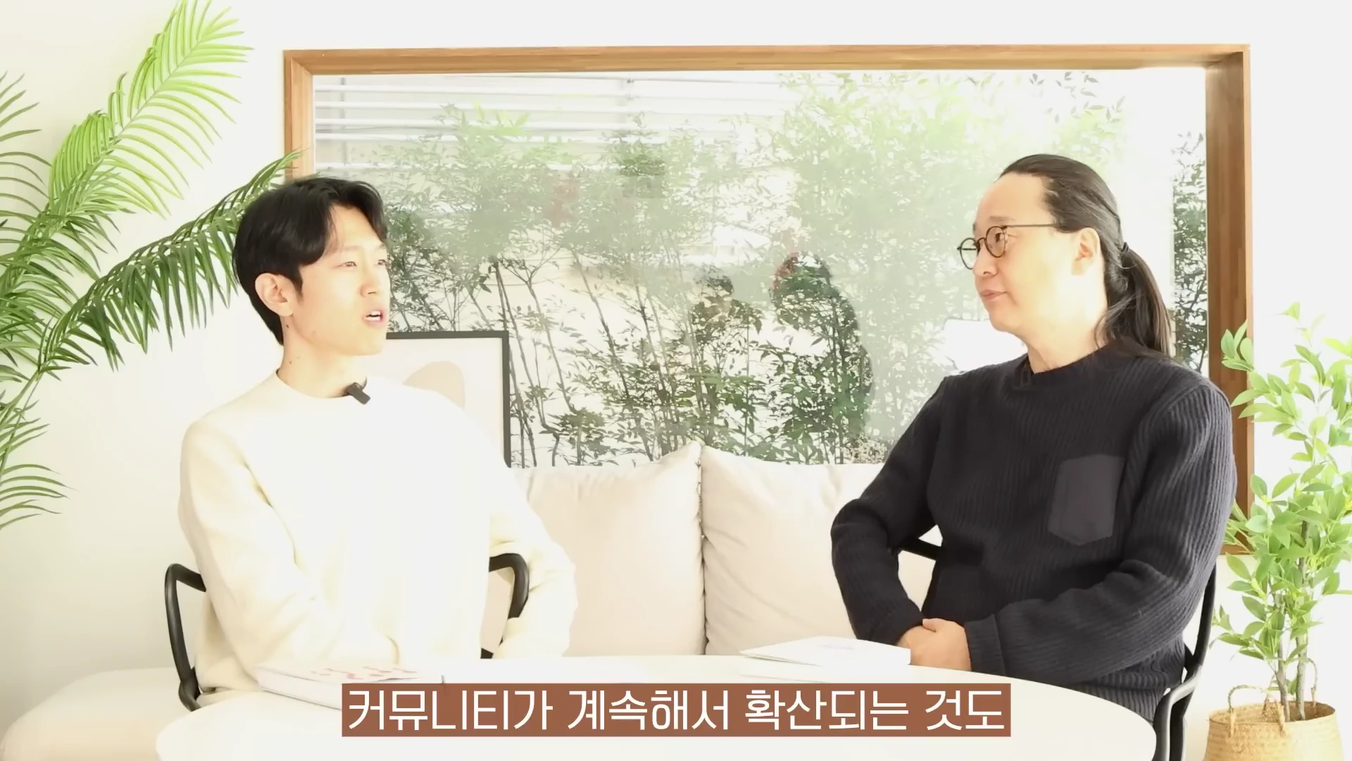 밝은 실내에서 두 남성이 마주 보고 앉아 대화를 나누고 있으며 화면 하단에는 자막이 나타나 있다.