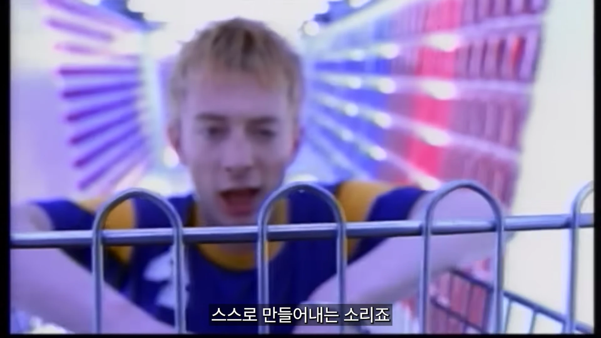 금발의 남성이 쇼핑 카트 너머로 카메라를 응시하며 노래를 부르고 있고, 배경은 복도와 같은 공간으로 흐릿하게 처리되어 있습니다.