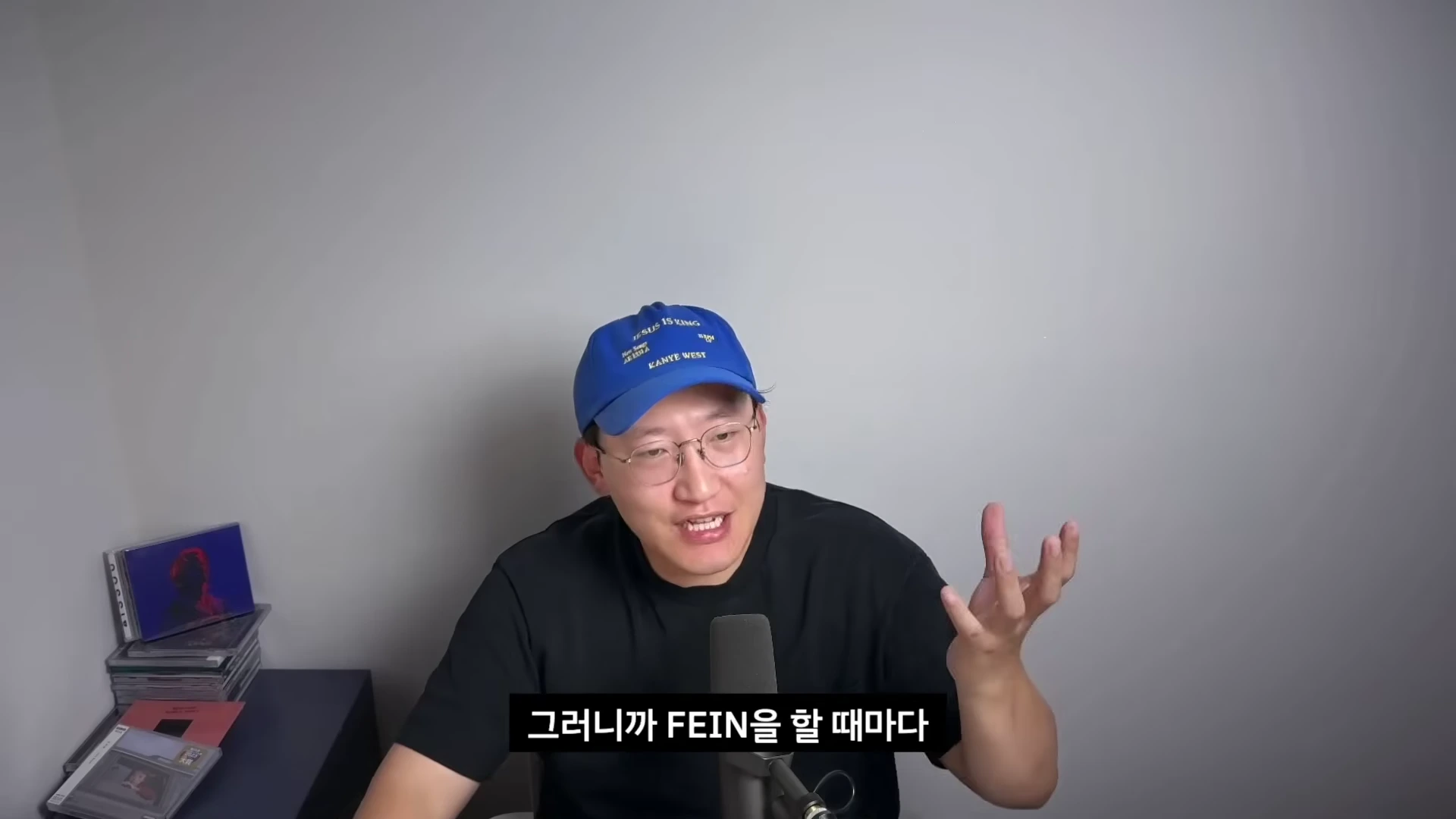 파란색 야구 모자를 쓴 안경을 낀 남성이 마이크 앞에서 손짓하며 무언가를 설명하고 있다.