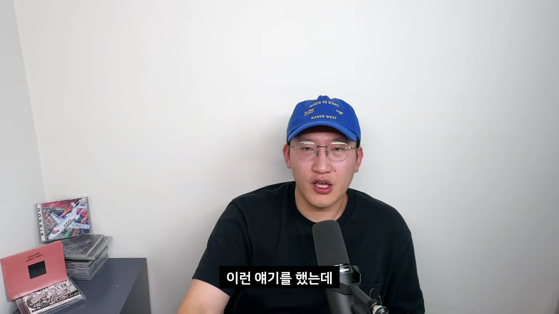 파란색 야구 모자와 안경을 쓴 남성이 마이크 앞에 앉아 정면을 바라보며 이야기하고 있다.