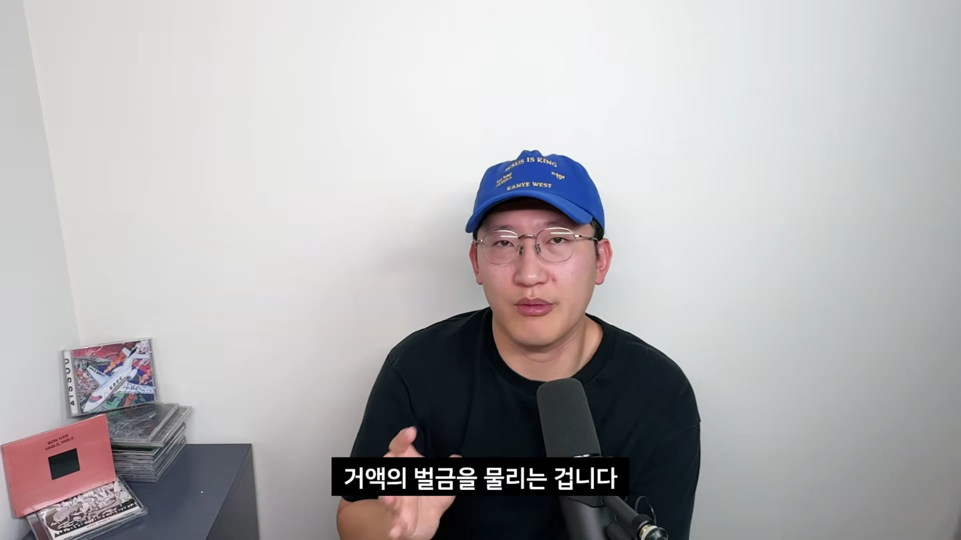 파란색 야구 모자와 안경을 쓴 남성이 마이크 앞에서 손동작을 하며 이야기하고 있으며 화면 하단에는 '거액의 벌금을 물리는 겁니다'라는 자막이 표시되어 있다.