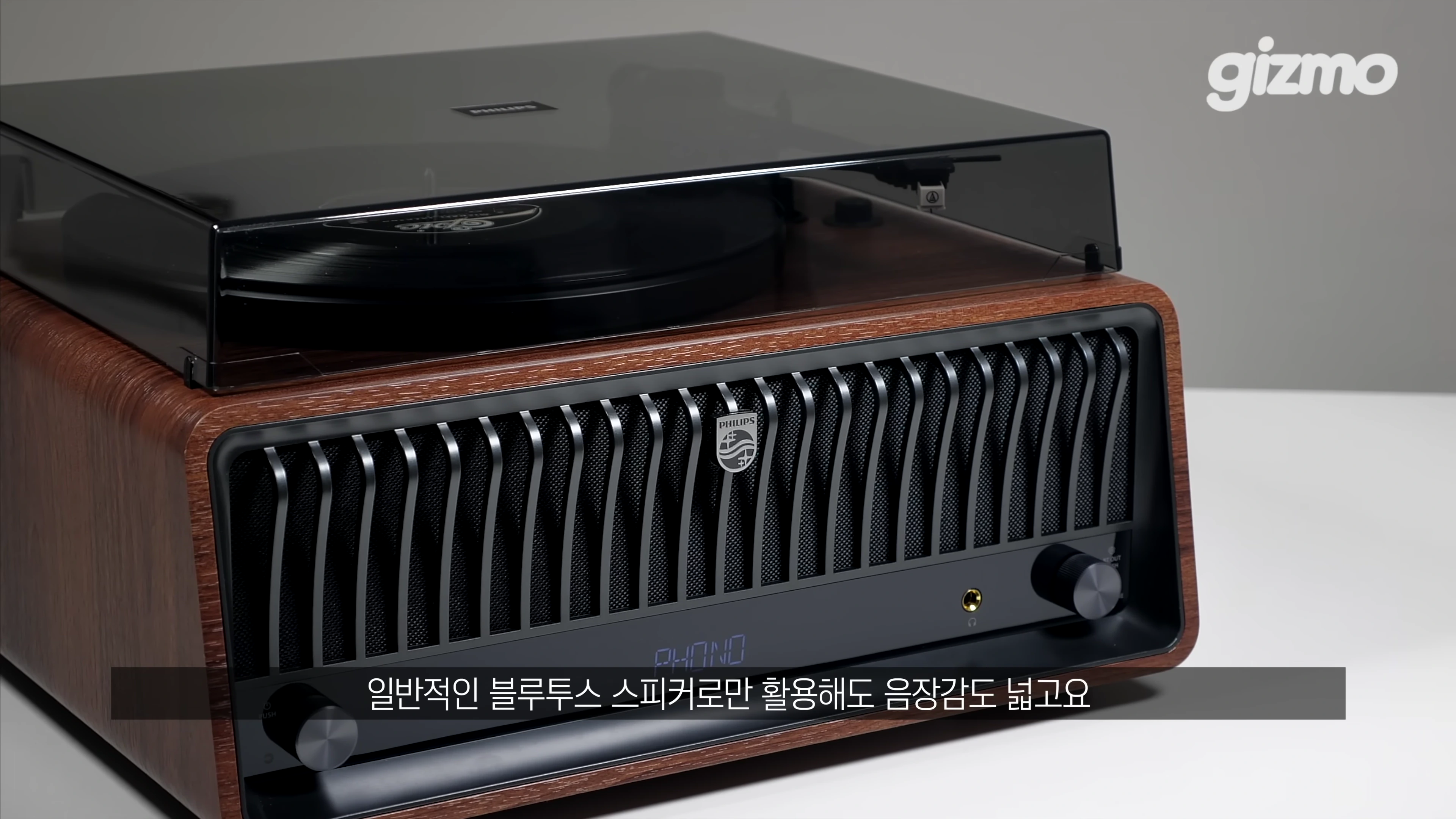 나무 질감의 외관을 가진 필립스 일체형 턴테이블이 밝은 배경 위에 놓여 있으며, 전면에는 스피커 그릴과 볼륨 다이얼, 디스플레이가 보입니다.
