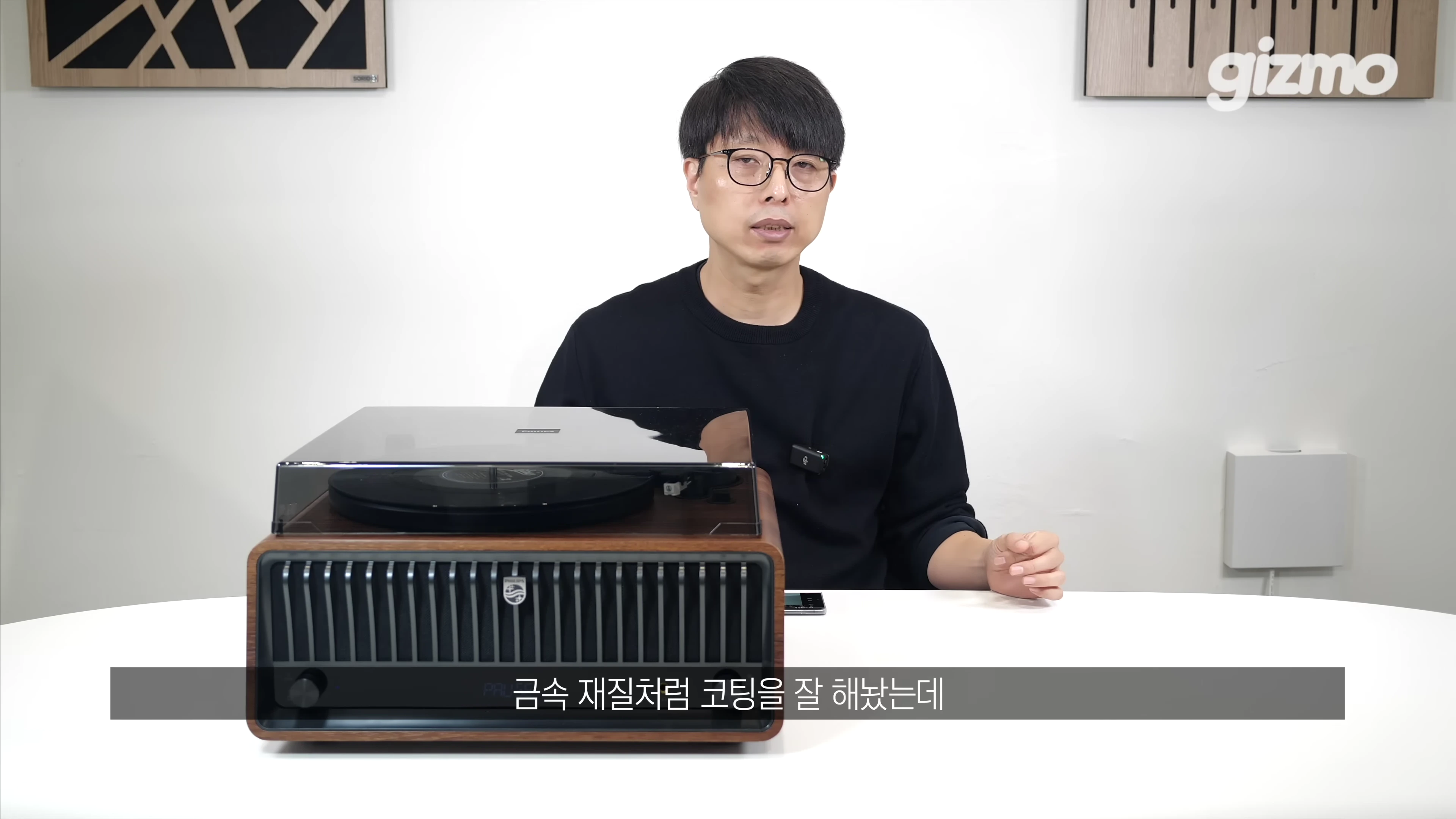 실내 스튜디오에서 한 남성이 테이블 위에 놓인 갈색 우드 톤의 필립스 일체형 턴테이블을 앞에 두고 설명하고 있다.