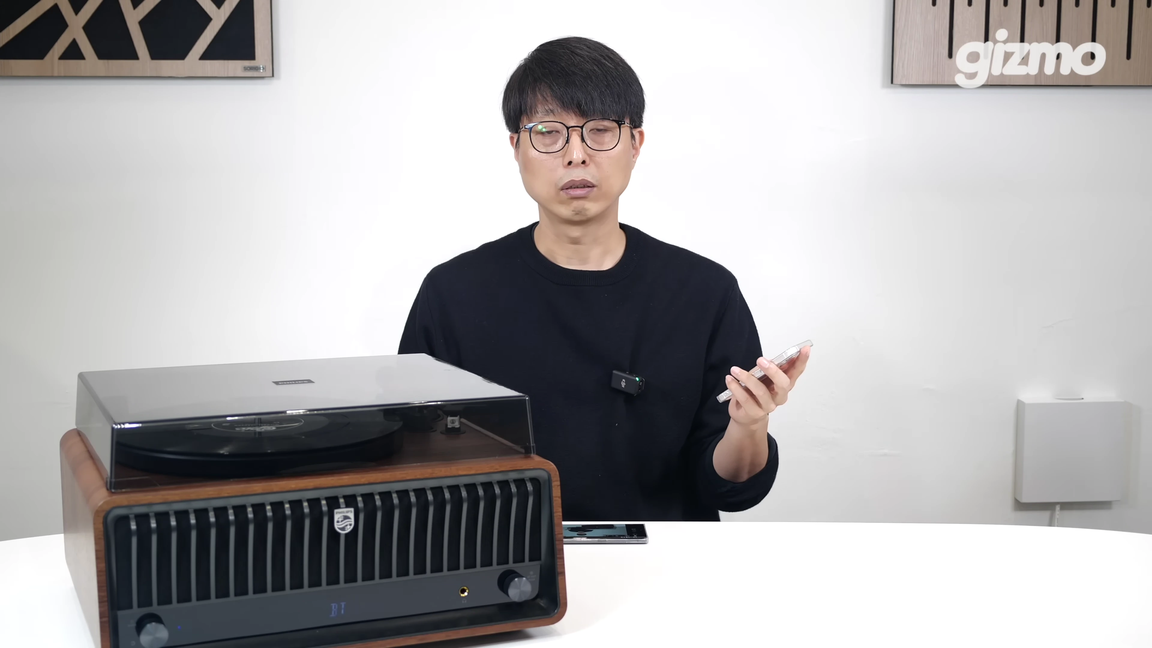 안경을 쓴 남성이 테이블 앞에 앉아 한 손에 스마트폰을 들고 설명하고 있으며, 그 옆에는 턴테이블이 놓여 있습니다.