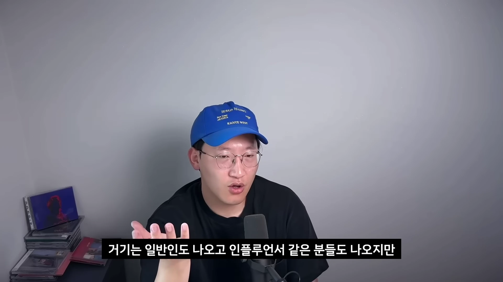 푸른색 야구 모자와 안경을 쓴 남성이 회색 벽을 배경으로 마이크 앞에서 이야기를 나누고 있다.
