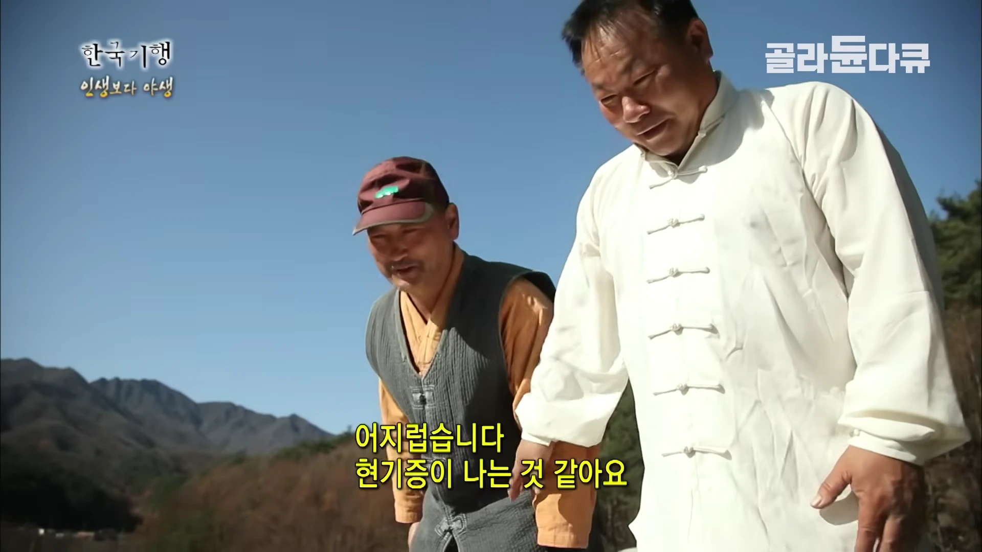 두 명의 중년 남성이 야외에서 기 수련을 하는 모습이다. 한 명은 흰색 무술복 차림이며, 다른 한 명은 조끼를 입고 캡 모자를 쓰고 있다.